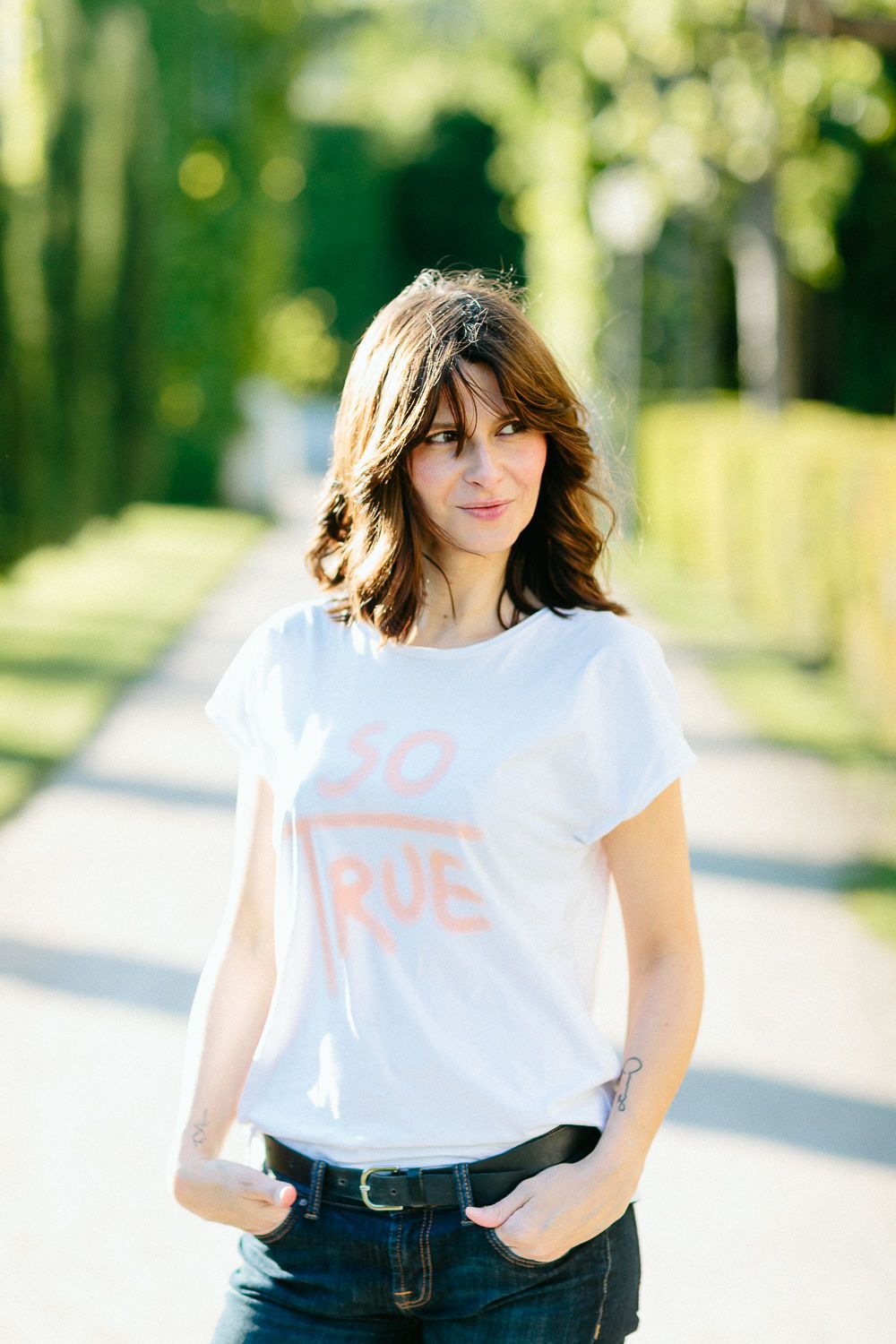 Brunette woman in white graphic t-shirt standing in sunny park pathway with hands in pockets.