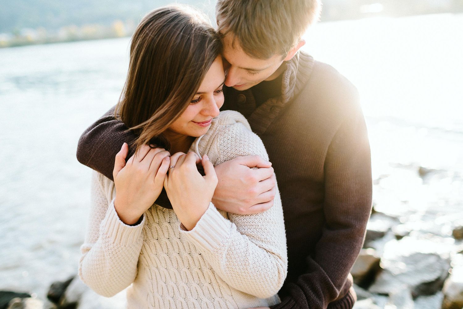 Couple embracing tenderly near a rocky shoreline with warm golden sunlight behind them.
