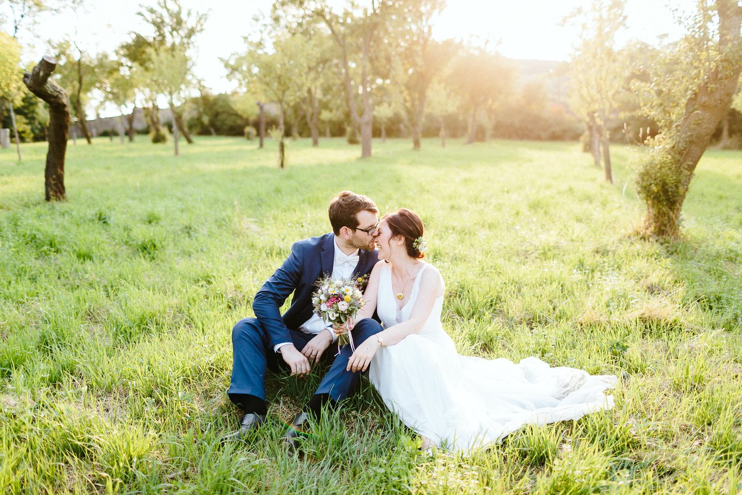 Bride and groom share a romantic kiss sitting in a sunny green orchard on their wedding day.