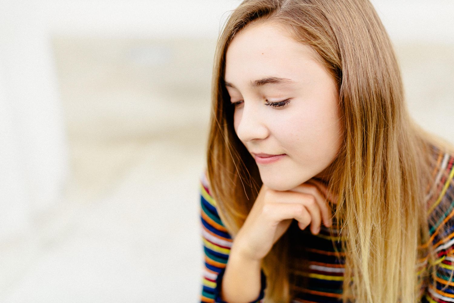Young girl with long blonde hair in a colorful striped sweater resting her chin on her hand thoughtfully.