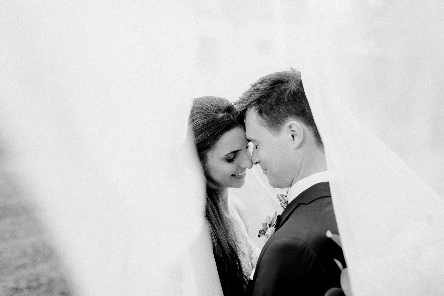 Black and white portrait of bride and groom touching foreheads beneath a flowing bridal veil.