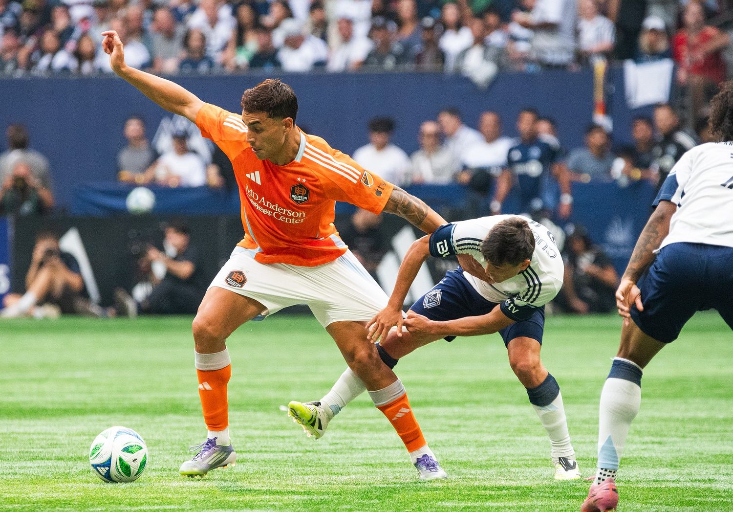 A Houston Dynamo soccer player in orange tries to kick the ball while a Vancouver Whitecaps player pulls the Houston player's shorts.