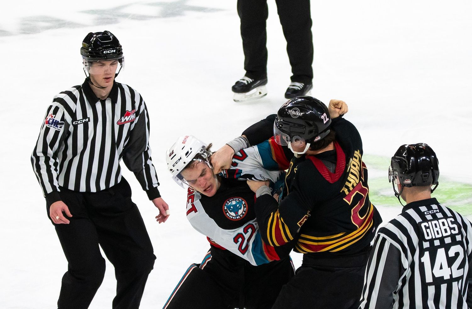 A Vancouver Giant hockey player in a black jersey punches a Kelowna Rocket hockey player in the face while the referees watch