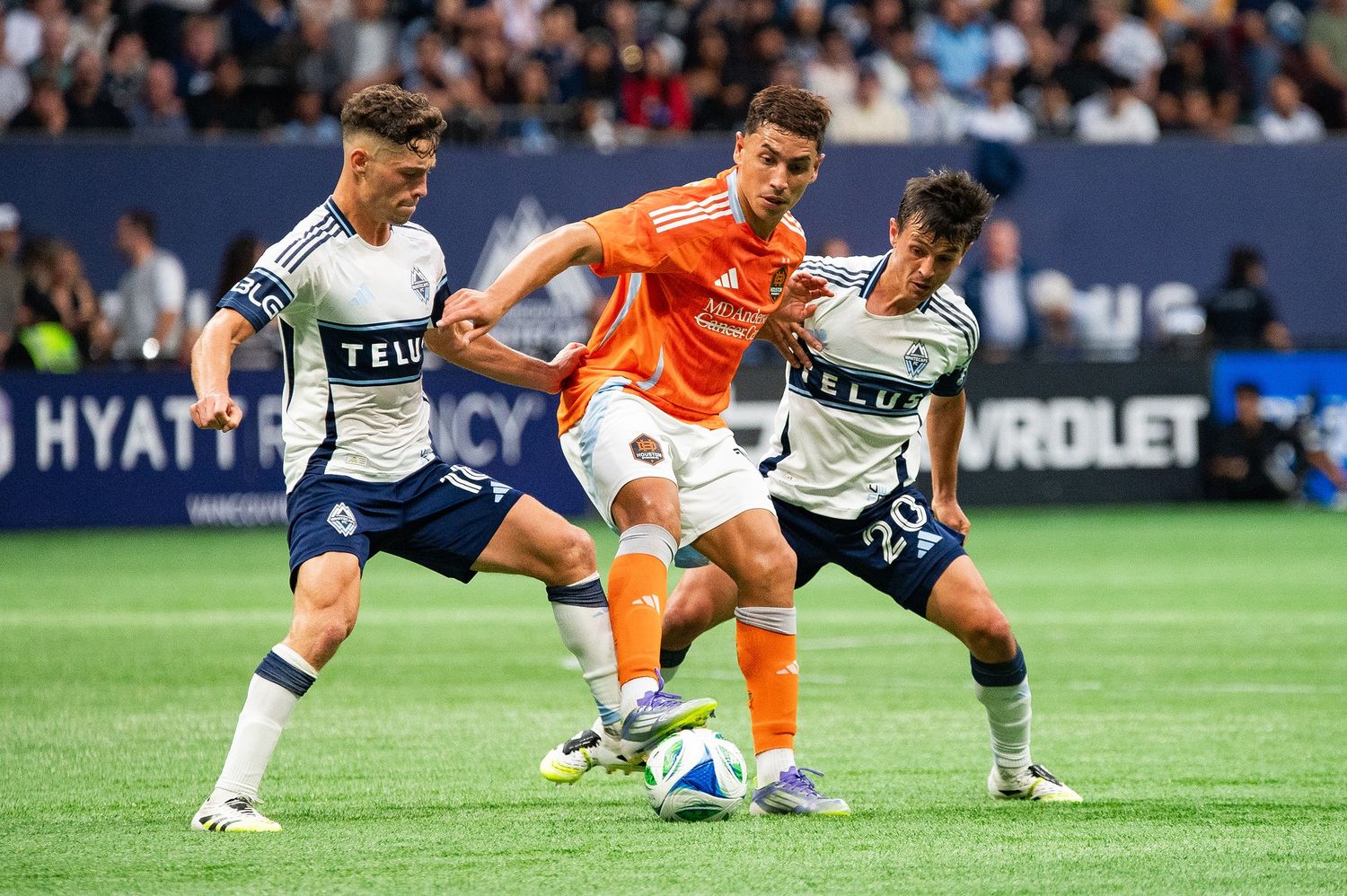 A Houston Dynamo soccer player in orange tries to keep the ball away from two Vancouver Whitecaps players