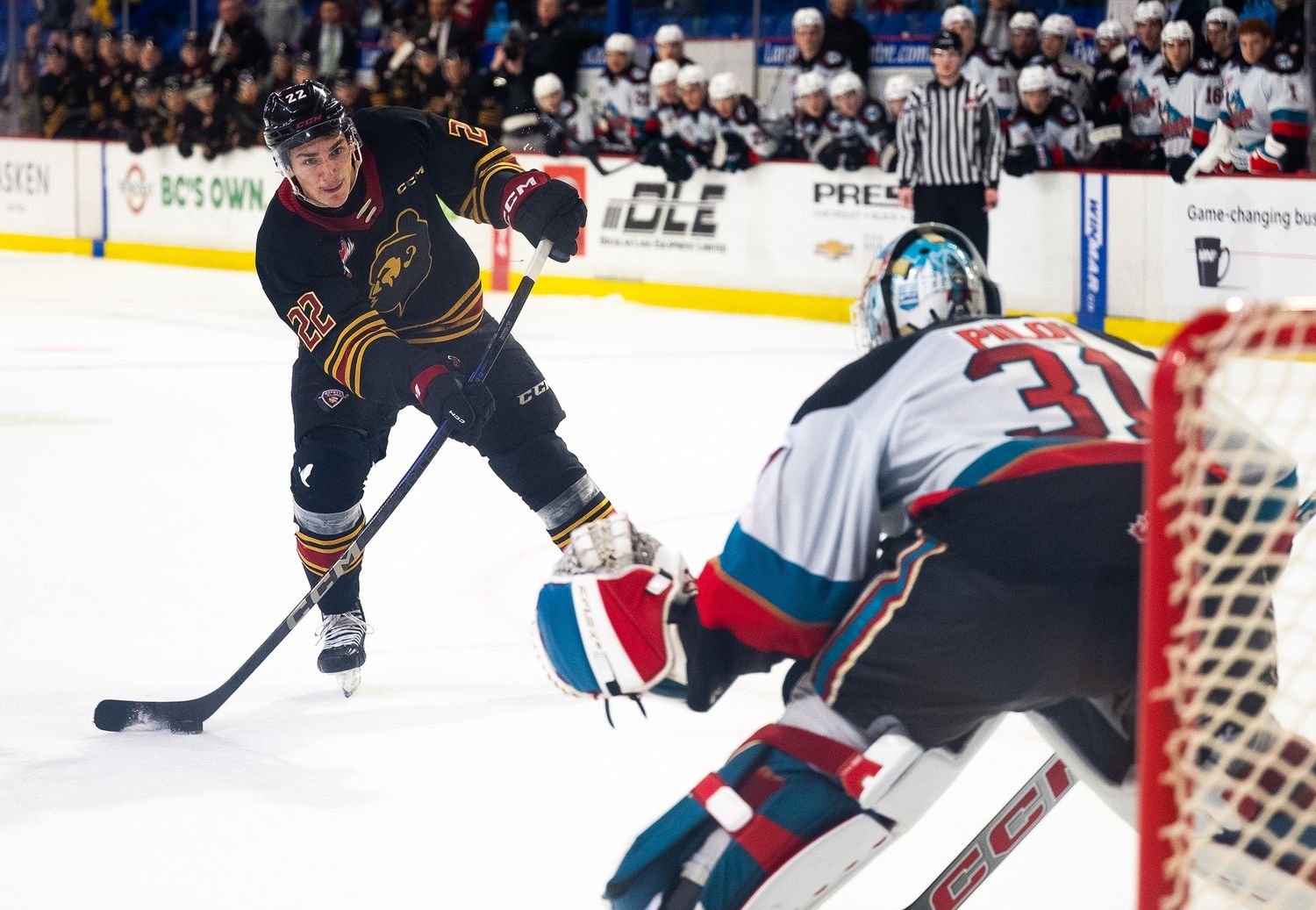 A Vancouver Giant hockey player is mid penalty shot against a Kelowna Rocket goalkeeper. Behind him on the bench all the players watch him.