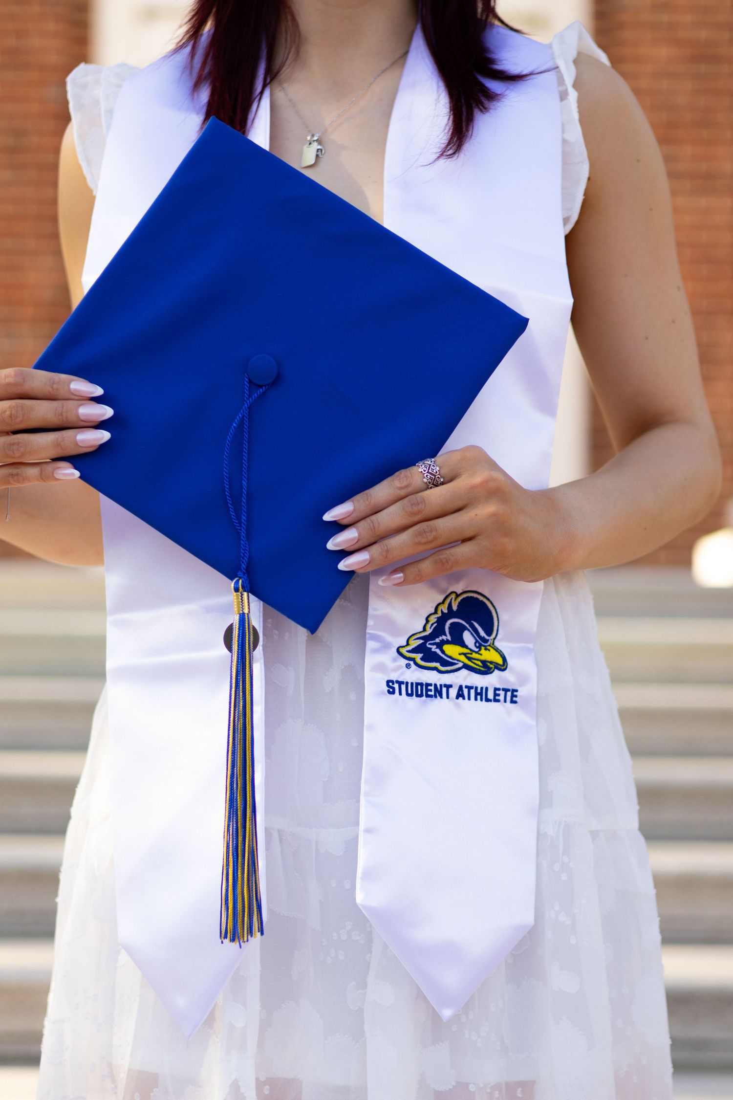 Graduate holding blue gradution cap with university logo and silver tassel against brick wall at the University of Delaware.