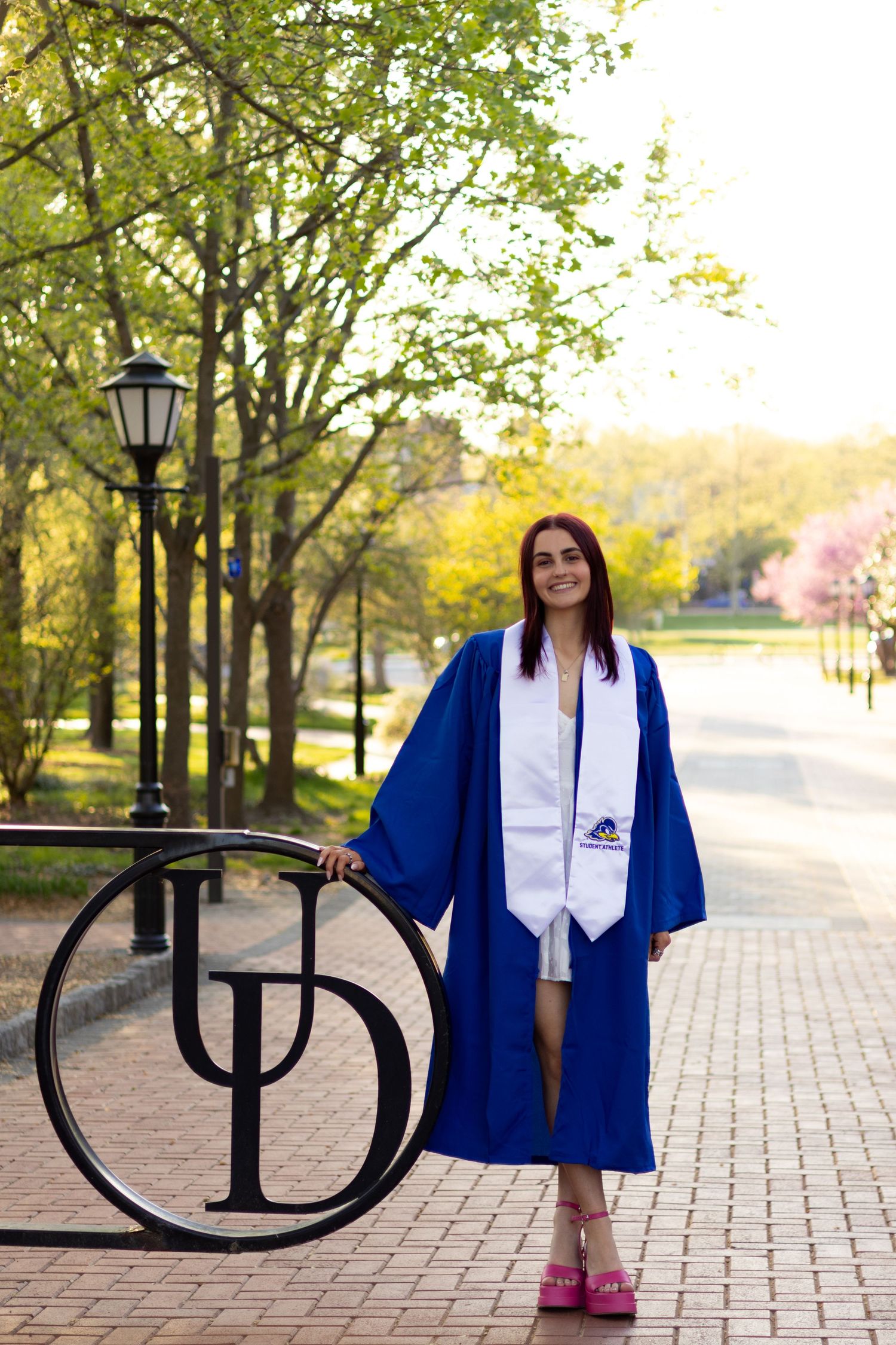 Graduation photos featuring blue cap and gown with decorative Greek letters on campus at the University of Delaware.