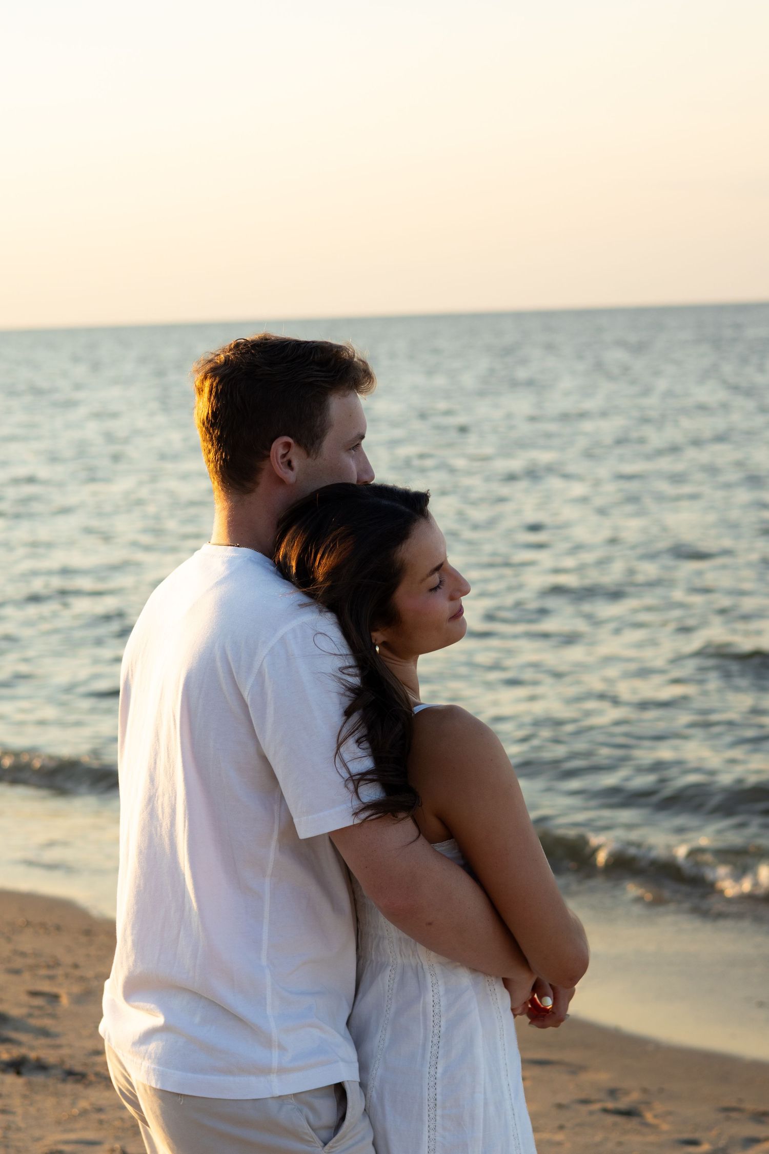 Couple embracing on beach at sunset wearing white clothing for engagement photos in Lewes, Delaware.