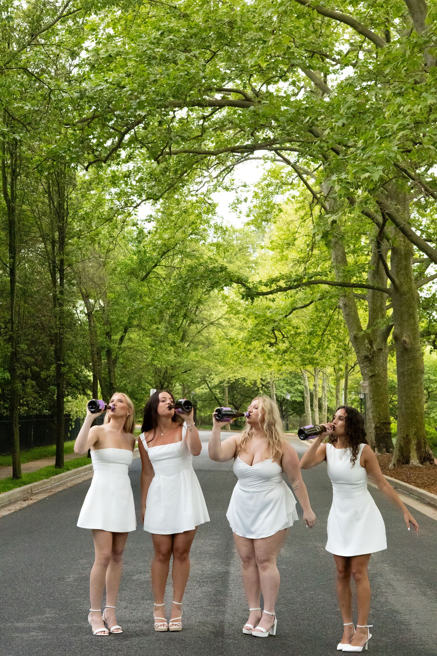 Graduates in white dresses walk away showing celebration on wooded campus path at the University of Delaware.