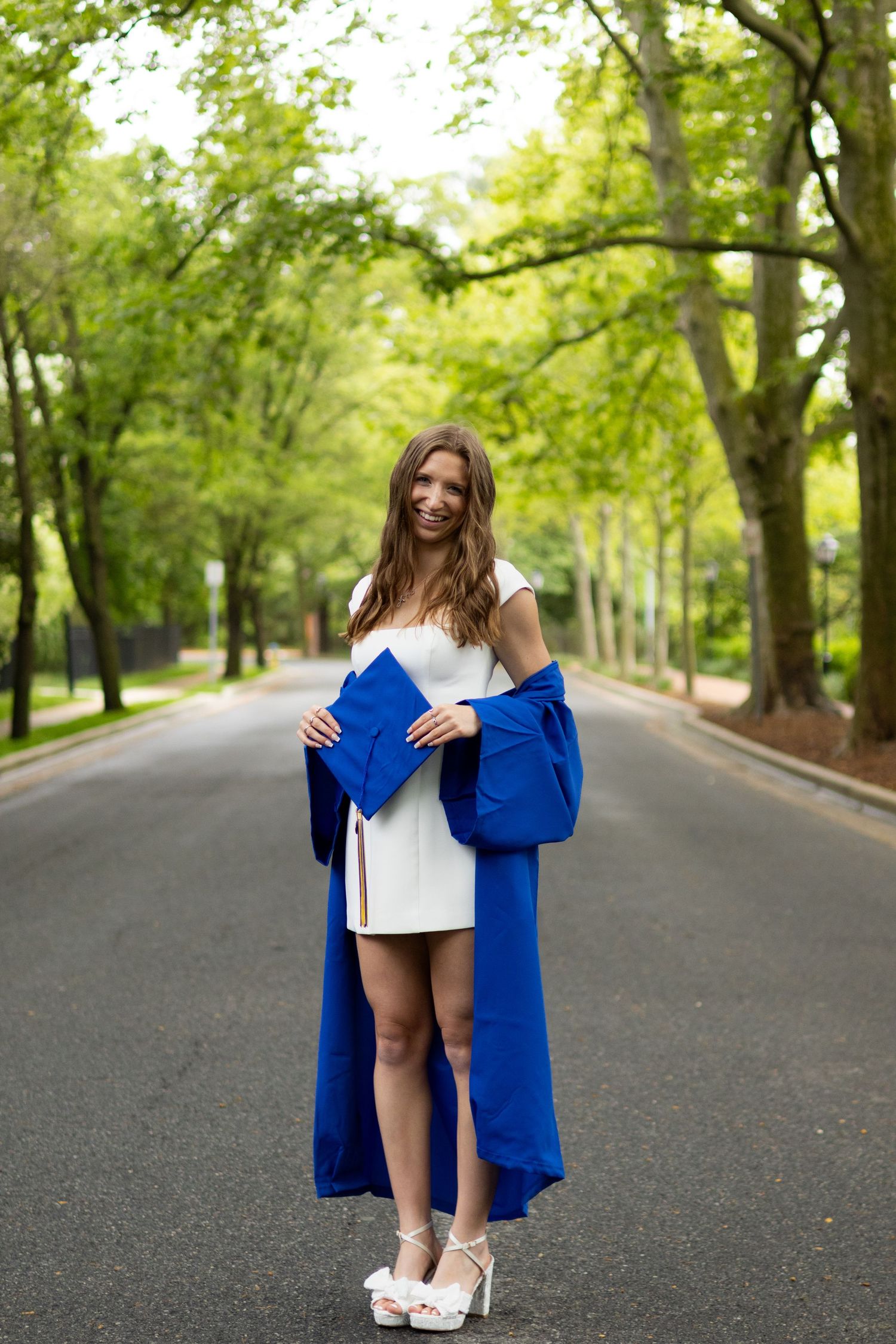 Graduate in blue cap and gown walking along a tree-lined path during outdoor graduation photo session at the University of Delaware.