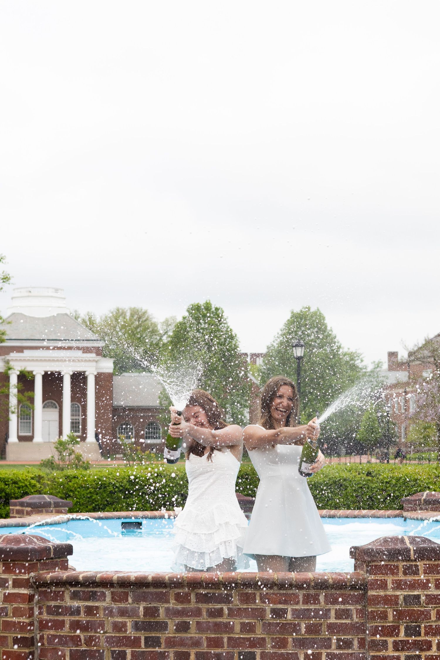 Two graduates in white dresses sit on brick wall with campus buildings in background at the University of Delaware.
