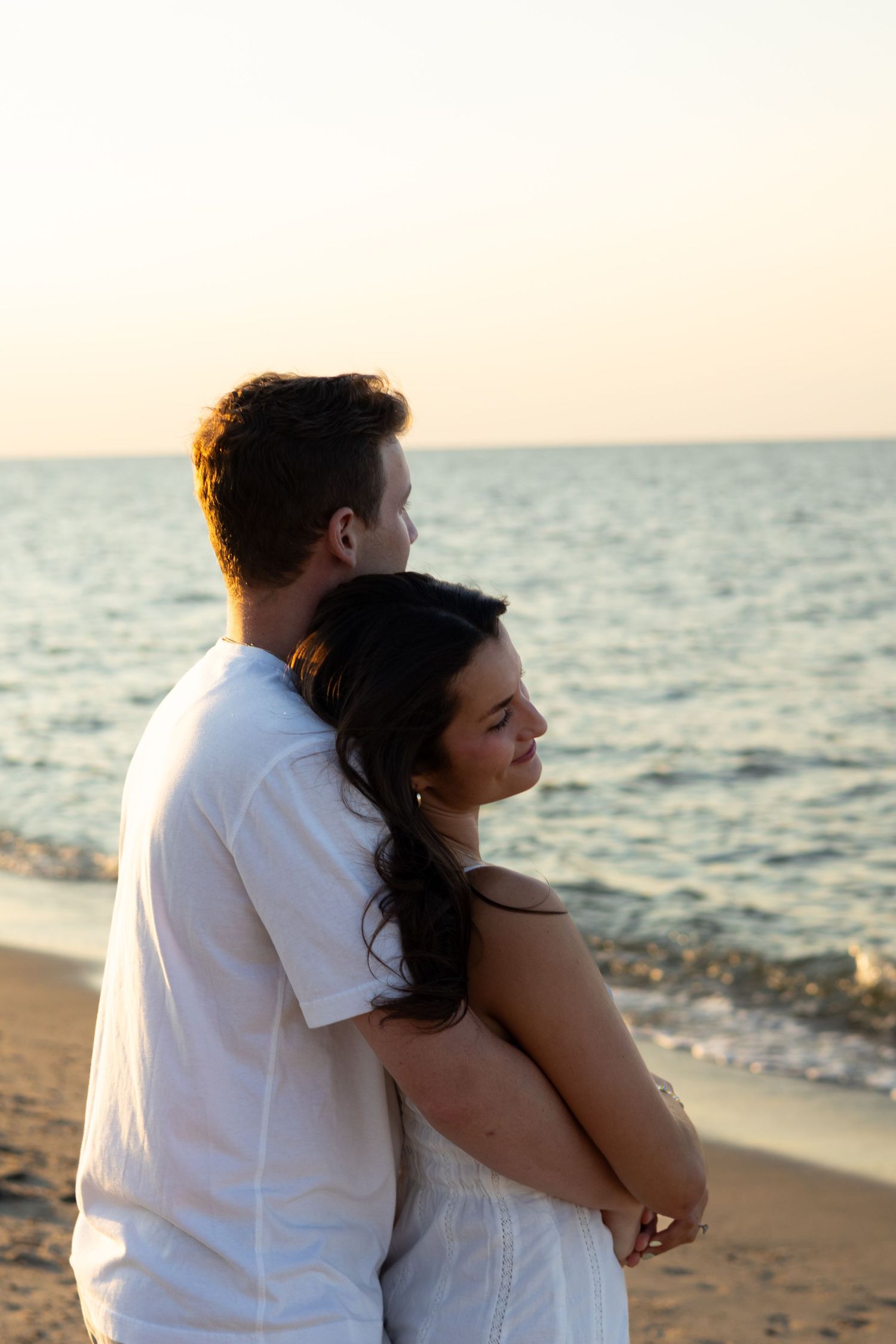 Couple embraces while watching sunset over ocean during beach engagement photoshoot in Lewes, Delaware.