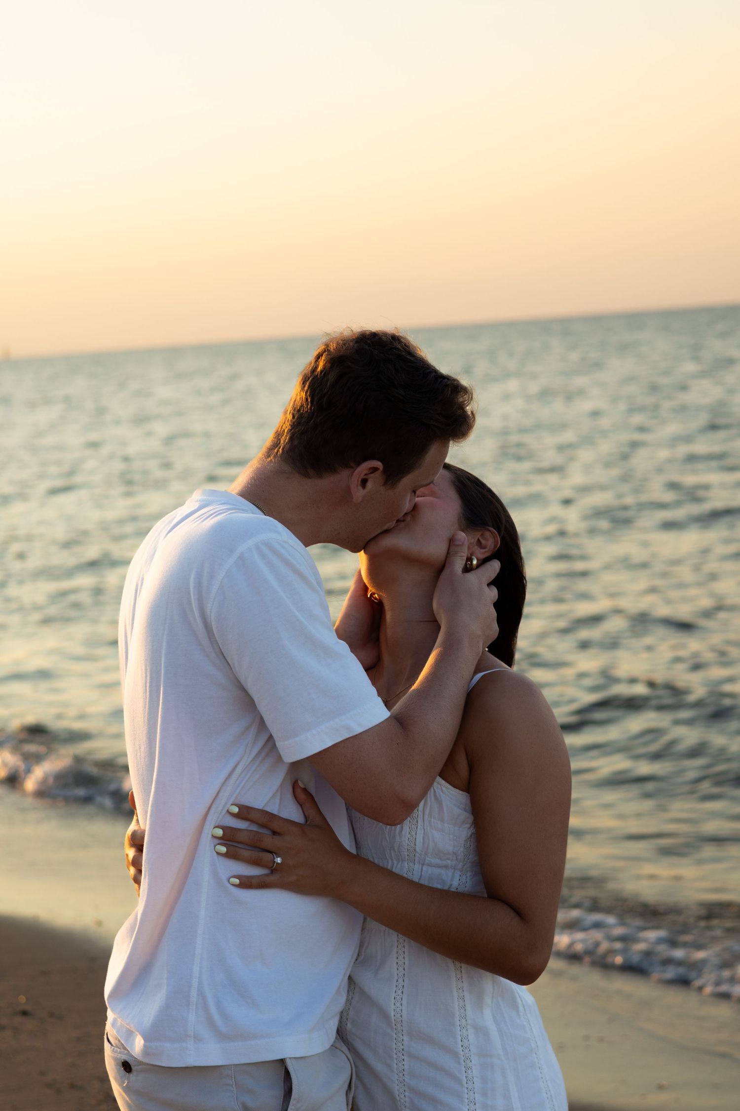 A romantic couple shares an intimate kiss on the beach at sunset with ocean waves in the background for engagement photos in Lewes, Delaware