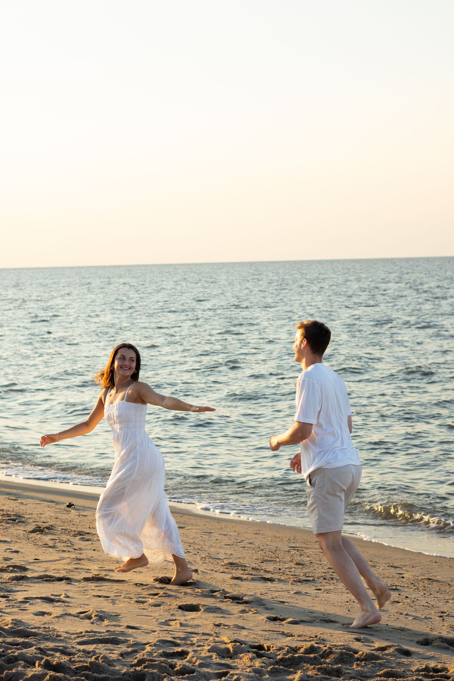 Couple in white clothing runs joyfully along the beach at sunset during engagement photos in Lewes Beach, Delaware.