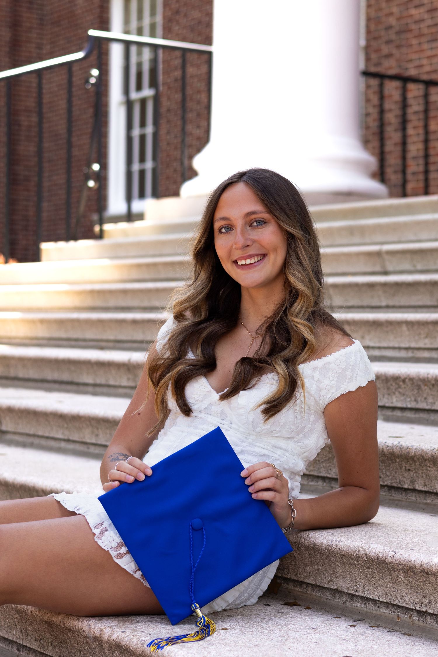 Graduate in white lace dress poses with graduation cap on stone steps at the University of Delaware.