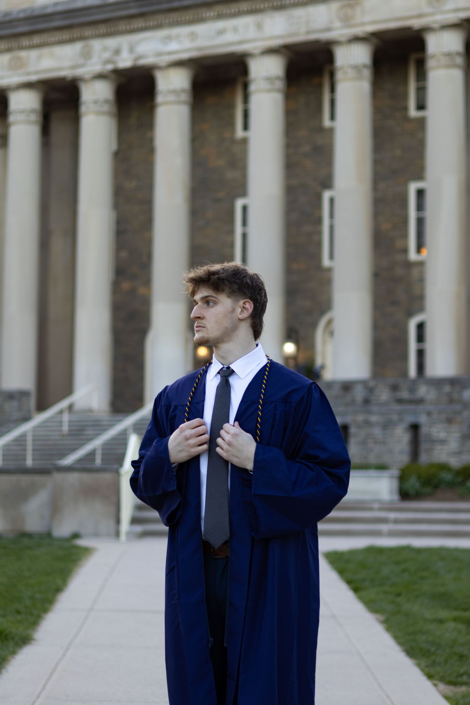 Graduate in navy blue cap and gown poses confidently in front of columned building facade at Penn State University.