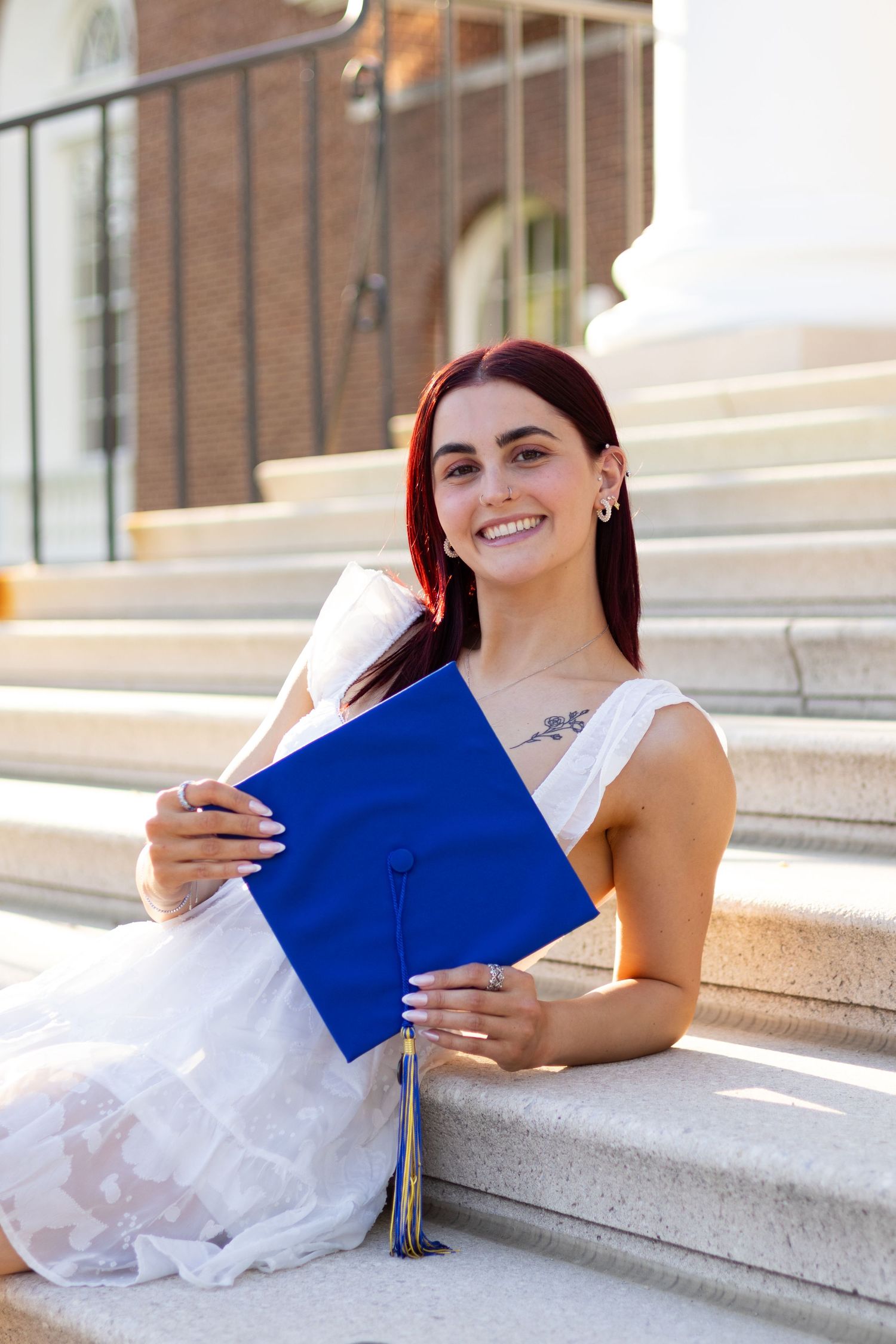 Graduate in white dress poses with blue graduation cap holder on concrete steps at the University of Delaware.