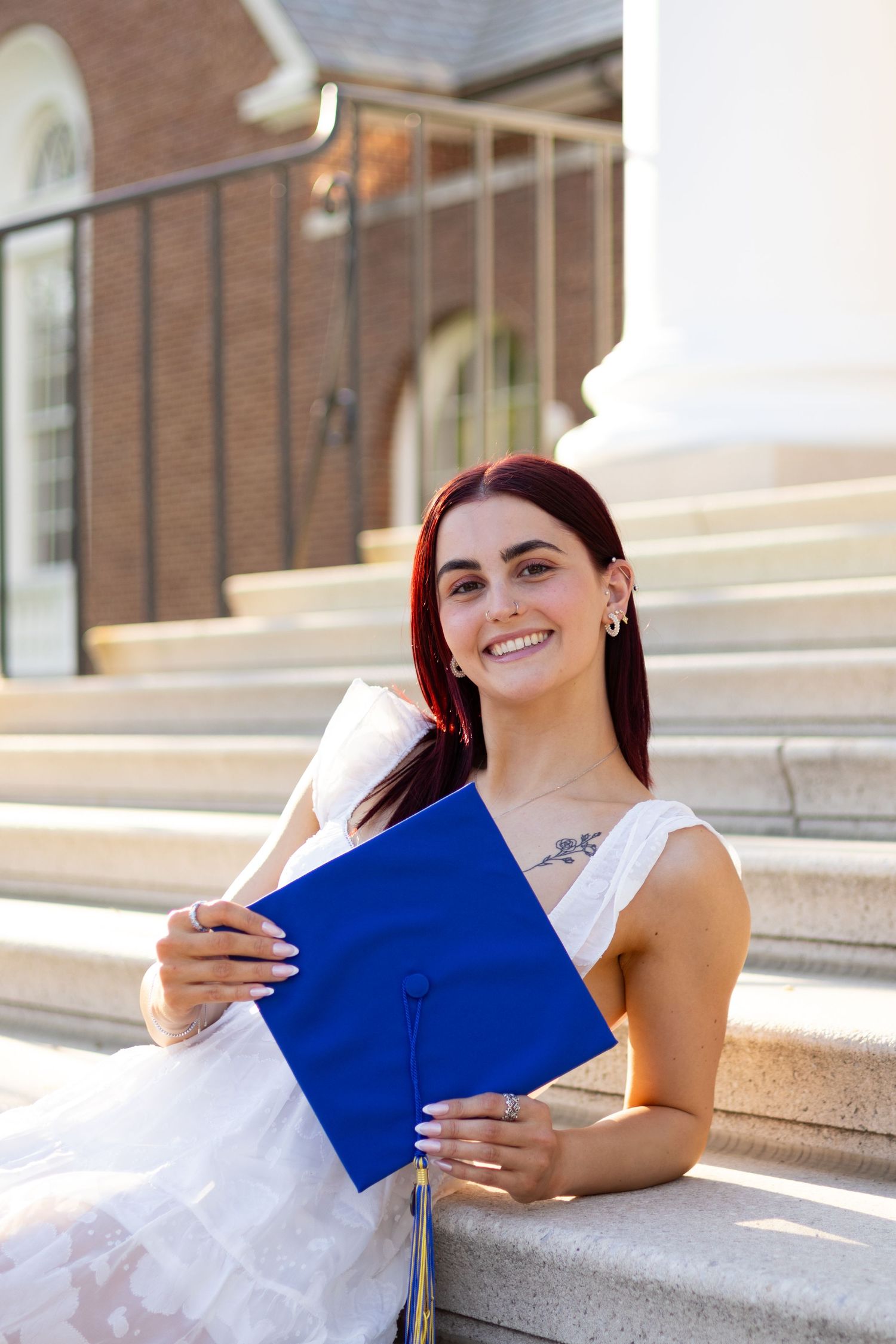 Graduate in white dress holding blue graduation cap on stone steps of academic building at the University of Delaware.