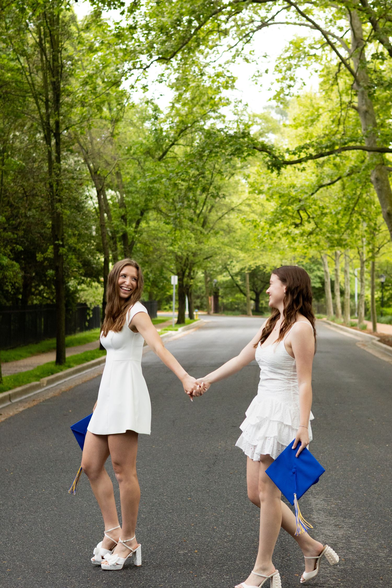 Two graduates in white dresses dance playfully on campus road at the University of Delaware.