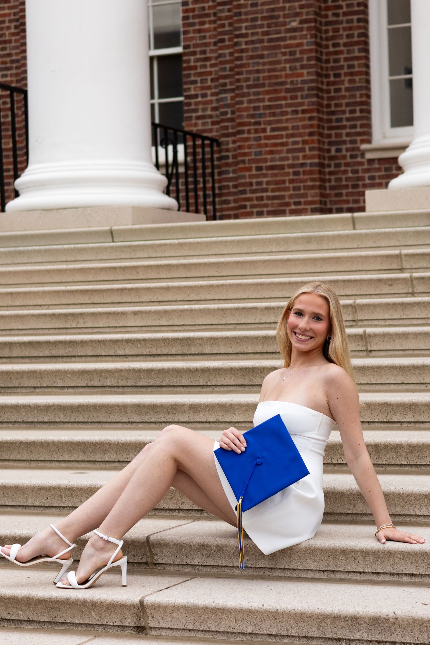 Graduate in white dress reclines on stone steps with blue graduation cap at the University of Delaware.