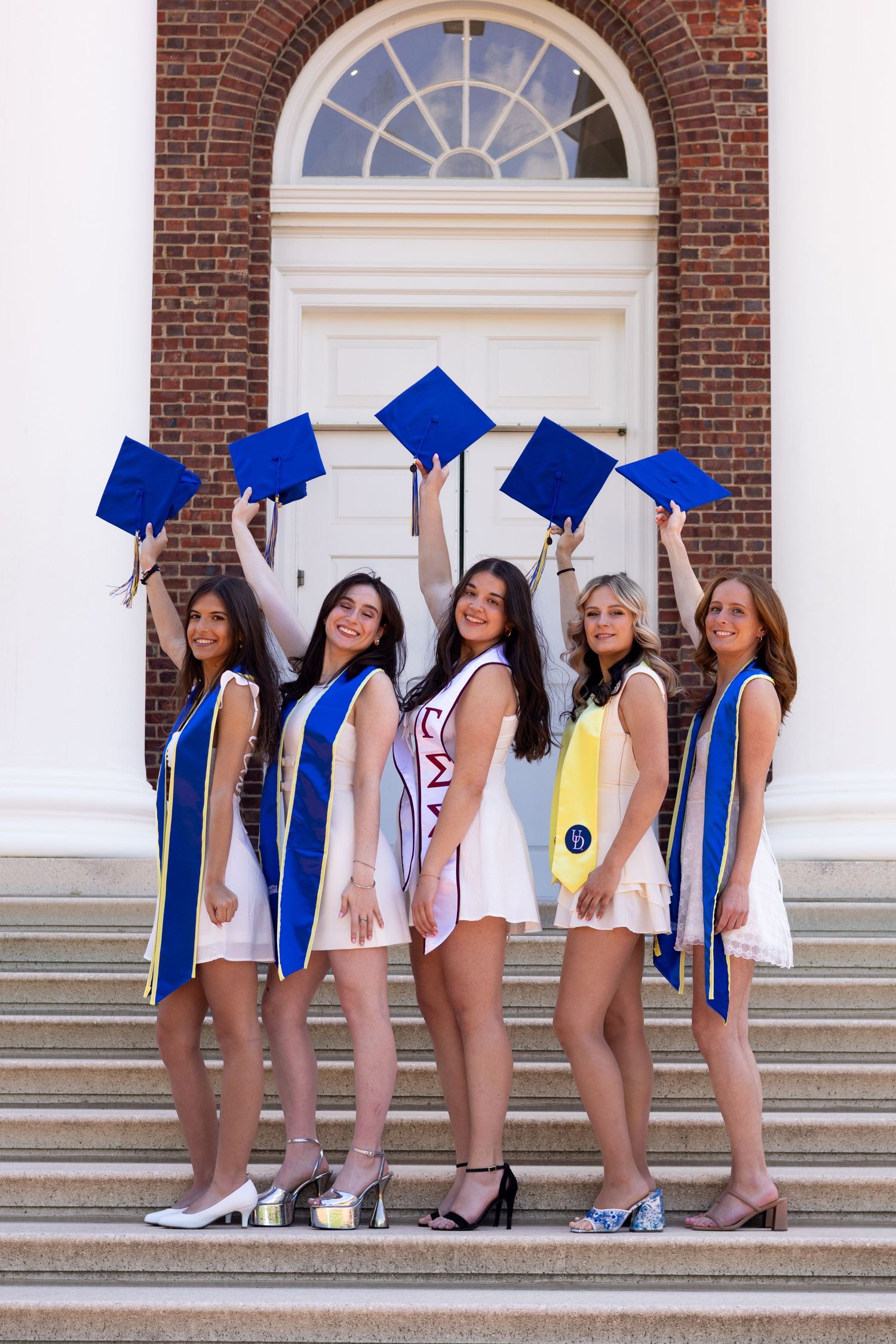 Graduates in white dresses pose with blue graduation caps on brick building steps at the University of Delaware.