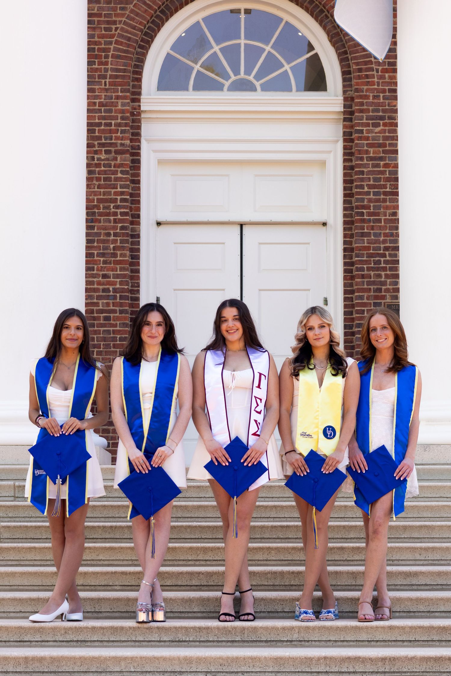 Group graduation photo with graduates in blue stoles sitting on brick building steps at the University of Delaware.