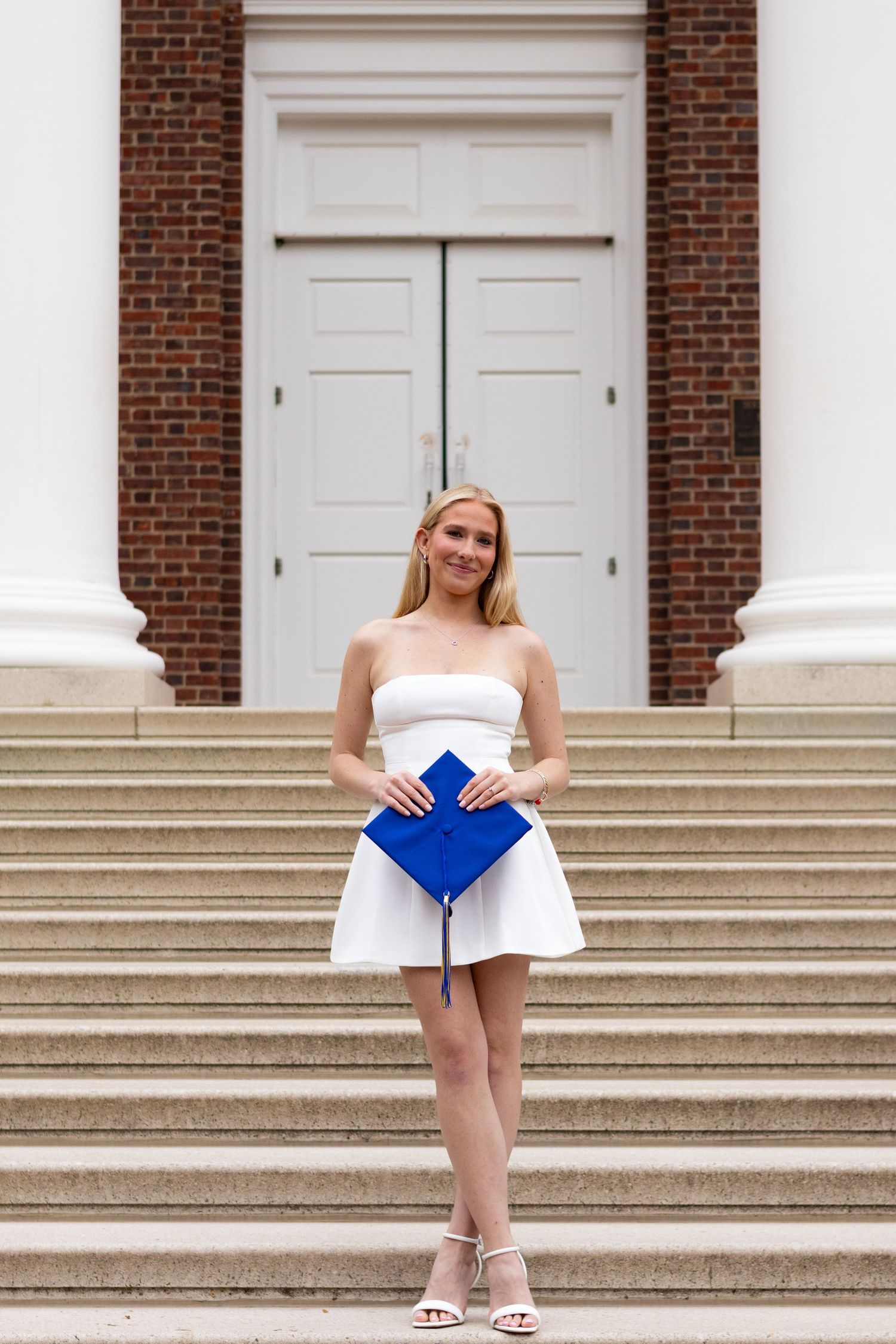 Graduate in white dress poses with blue graduation cap on stone steps at the University of Delaware.