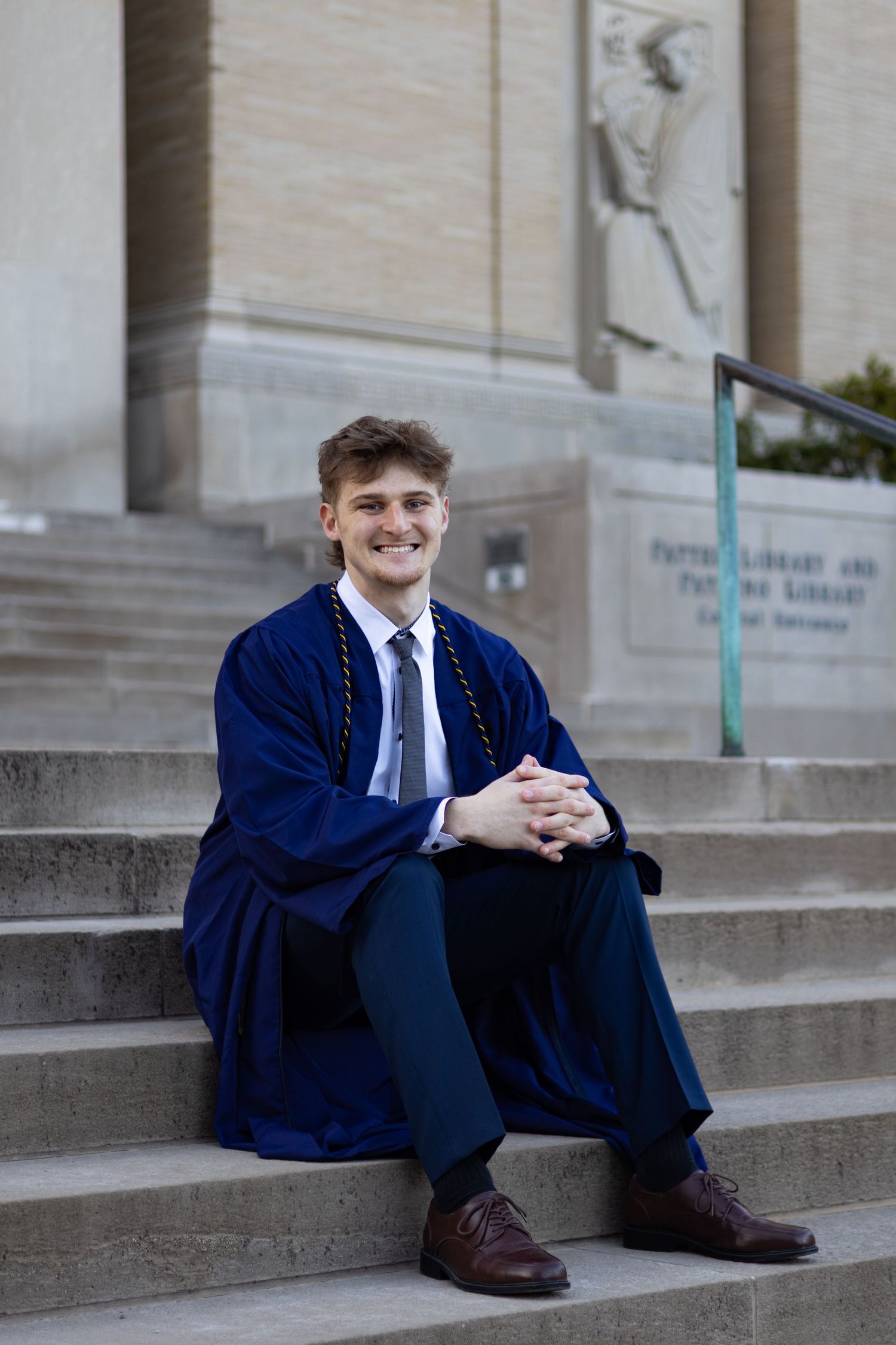 Graduate in blue academic regalia sits on concrete steps of campus building at Penn State University.