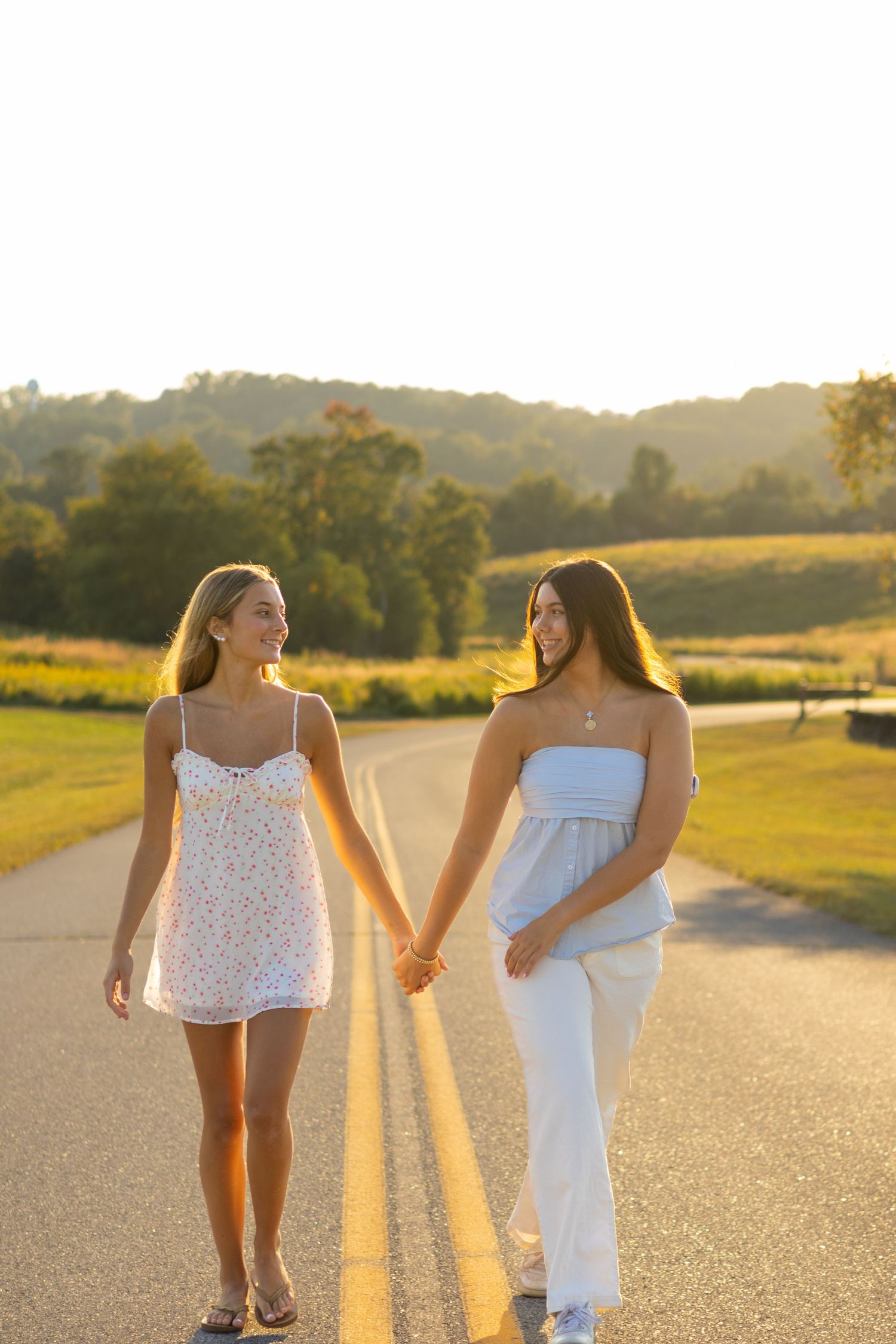 Two people in white outfits walk hand in hand on a sun-drenched road for senior photos in Wilmington, Delaware.