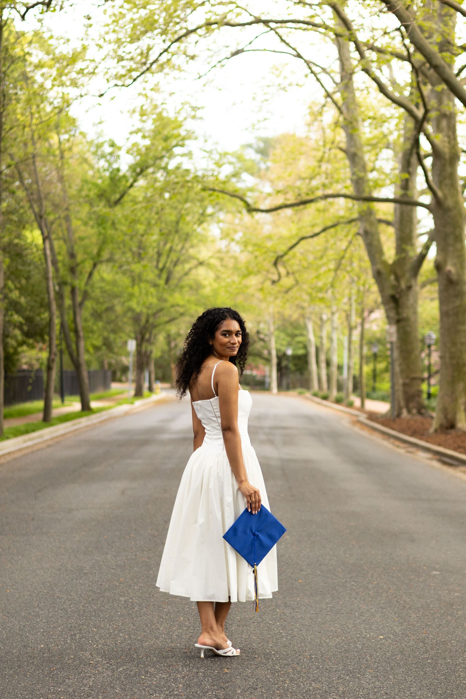 A white dress photoshoot on a tree-lined path with a blue graduation cap at the University of Delaware.