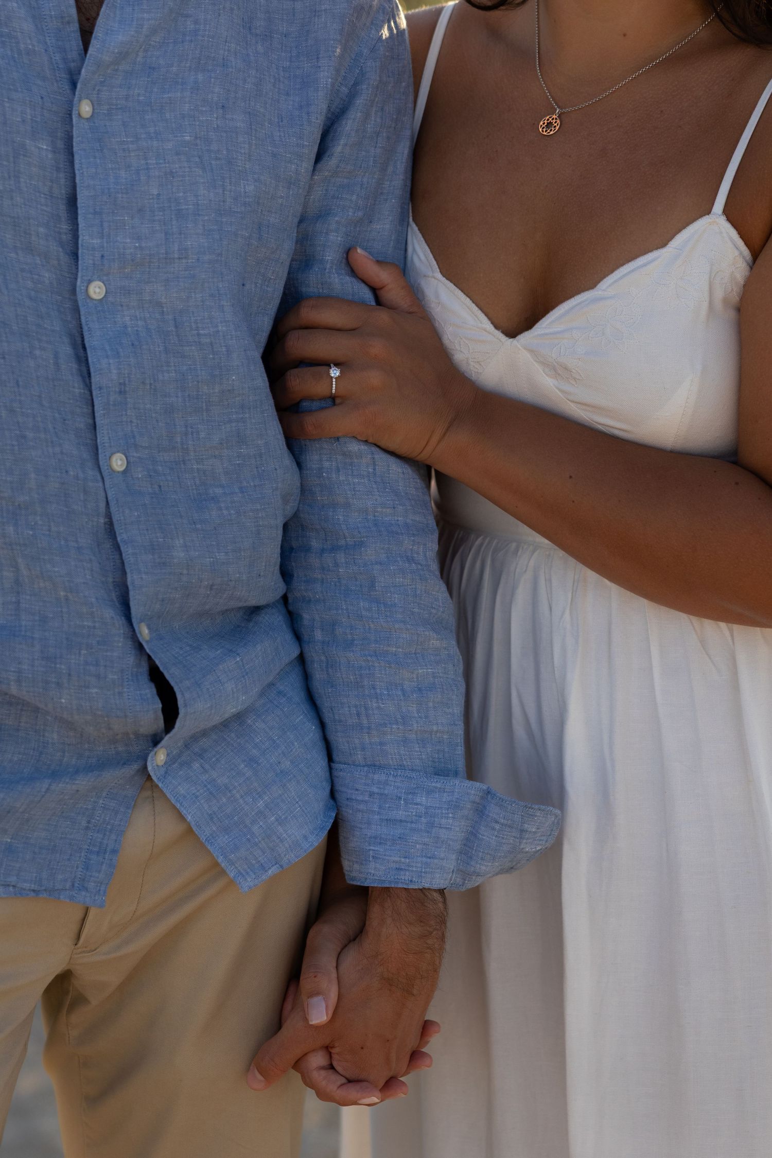 Close-up of couple in blue shirt and white dress holding hands with engagement ring visible In Lewes, Delaware.