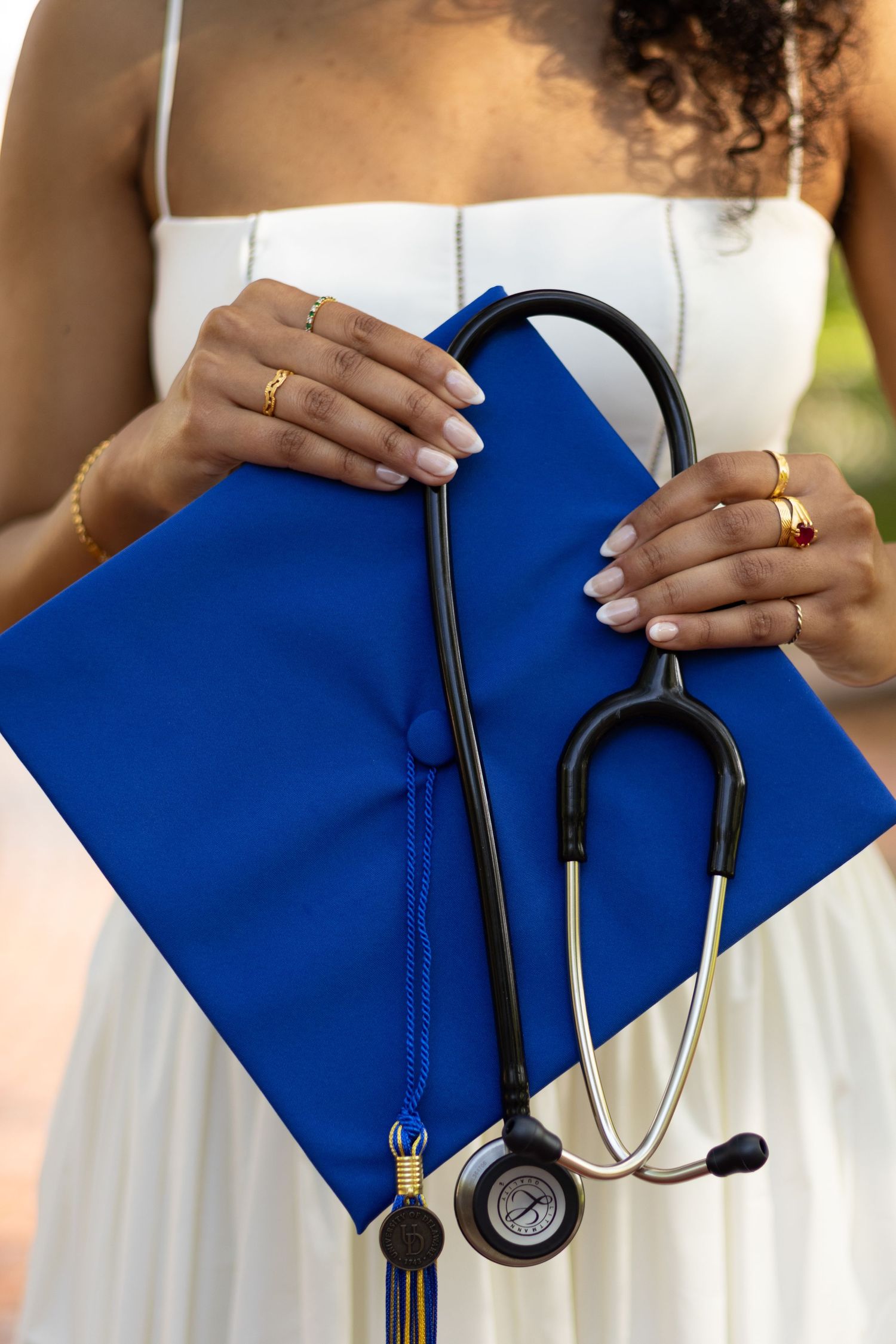 Close-up of hands holding a blue graduation cap and stethoscope against a white dress at the University of Delaware.