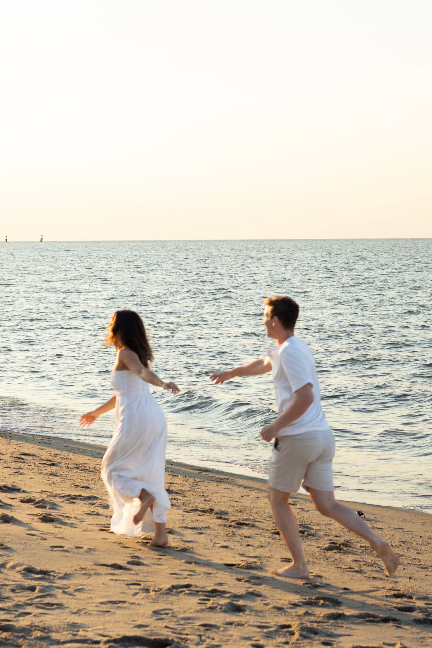 Couple chasing each other on sandy beach during golden hour for engagement photos in Lewes, Delaware.