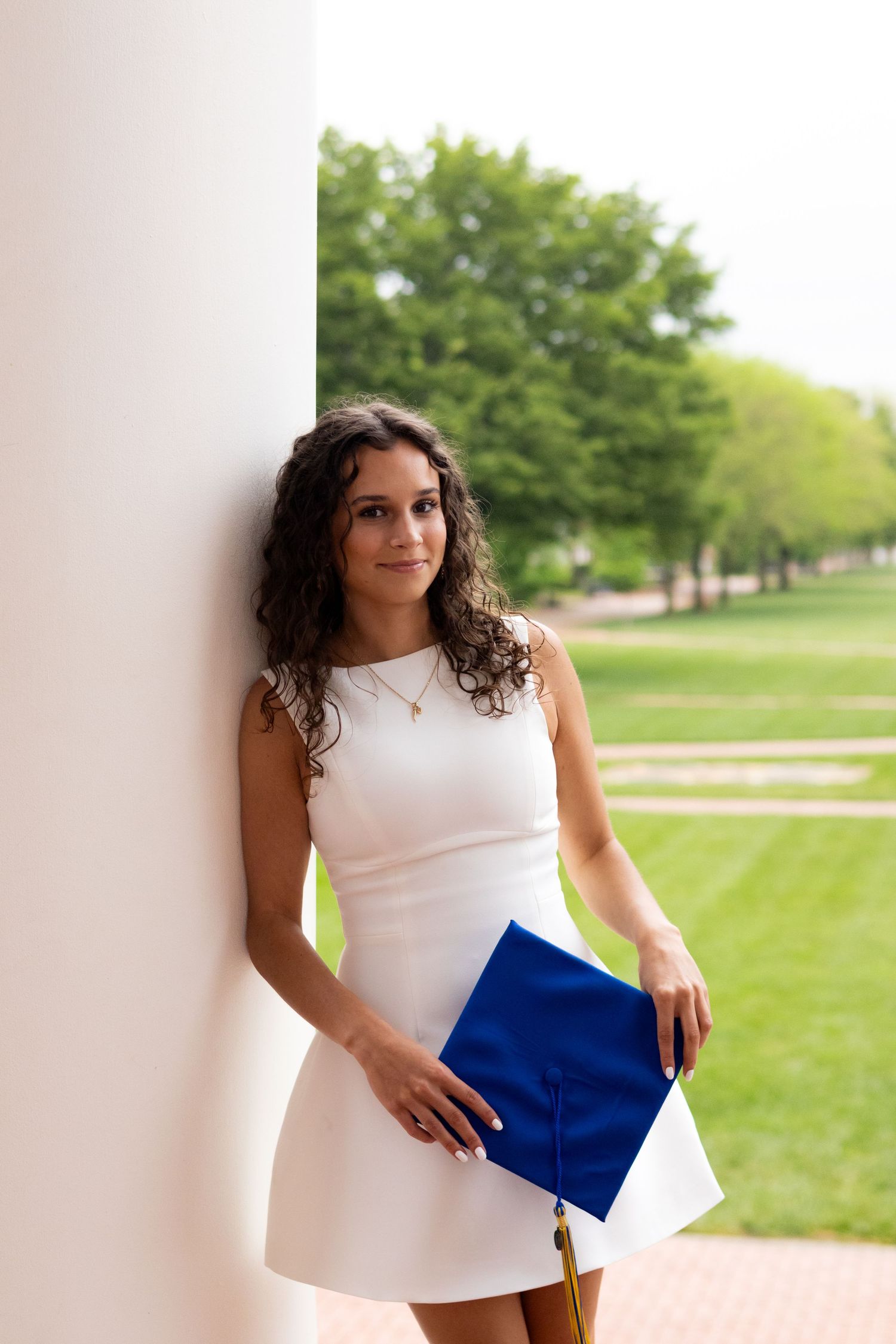 Graduate in white dress poses with blue diploma cover outdoors at the University of Delaware.