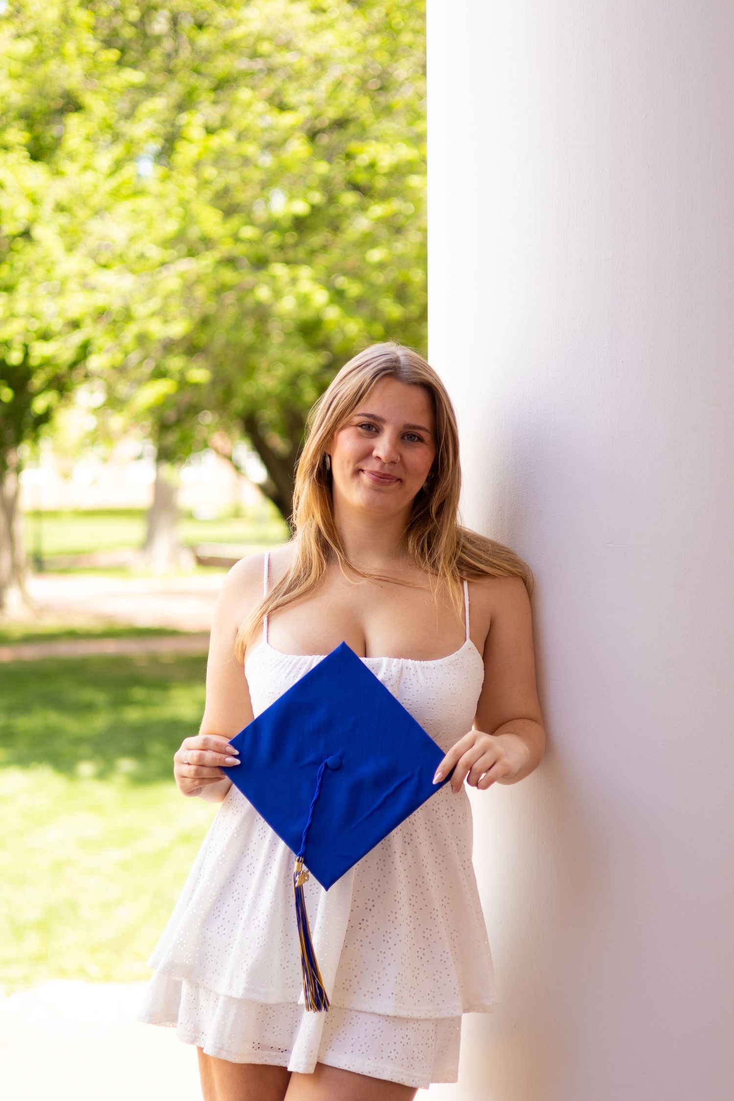 Graduate in white eyelet dress poses with blue graduation cap against white columns at the University of Delaware.