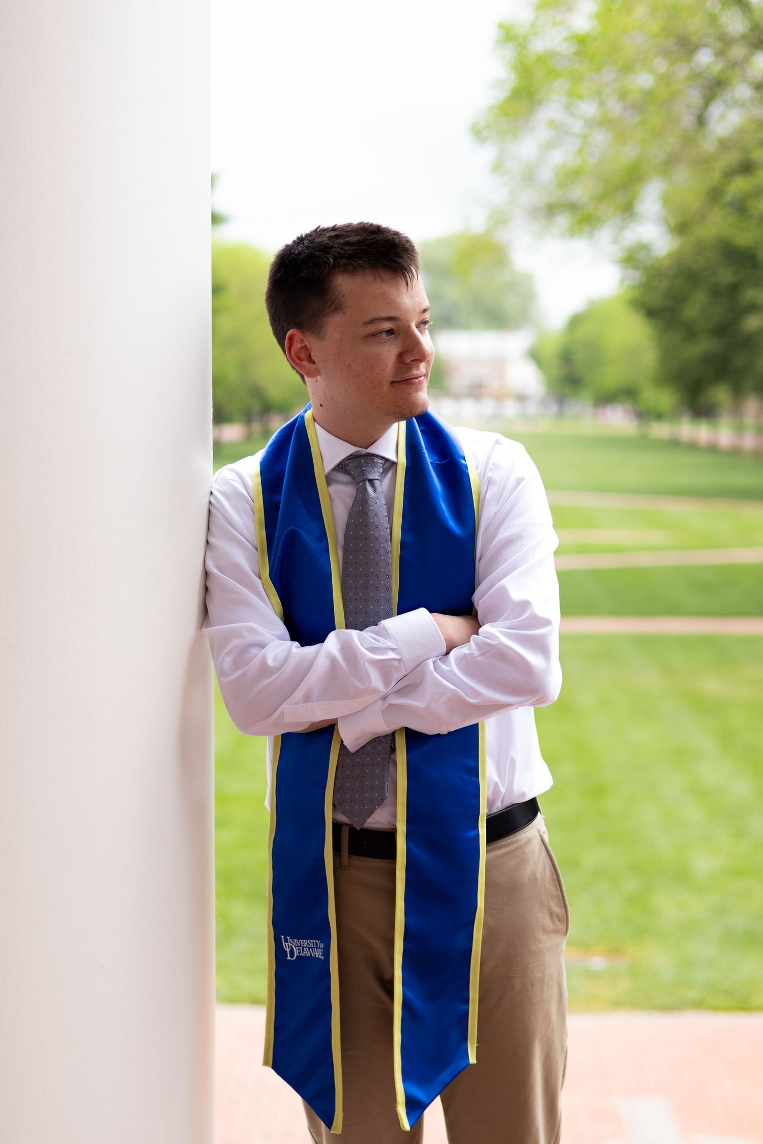 Graduate wearing blue stole and khaki vest poses against white column outdoors at the University of Delaware.