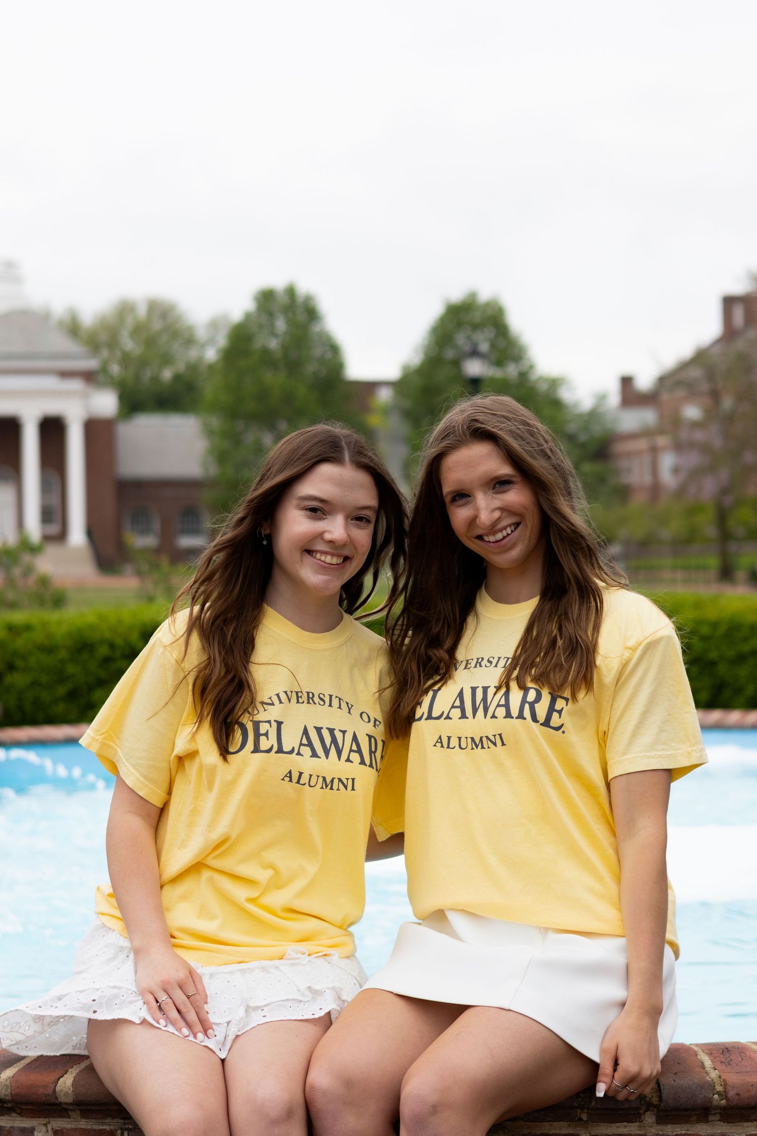 Two graduates wearing yellow Delaware alumni shirts sit together outdoors at the University of Delaware.