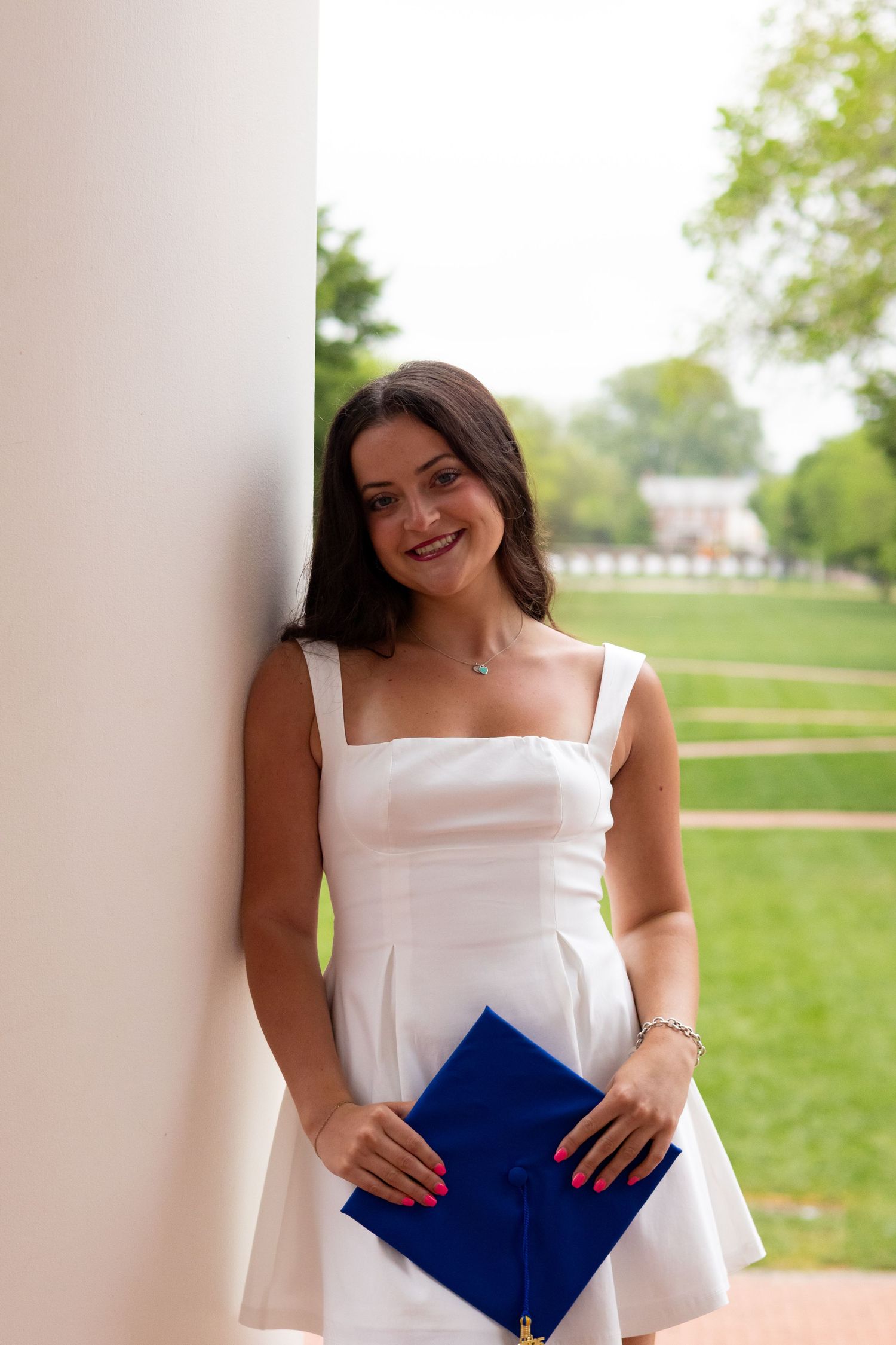 Graduate in white dress holds blue graduation cap while posing on campus at the University of Delaware.