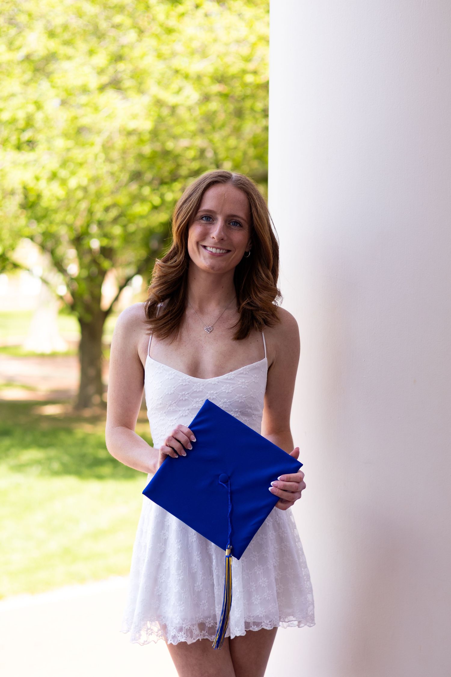 Graduate in white lace top poses with blue graduation cap by white columns at the University of Delaware.