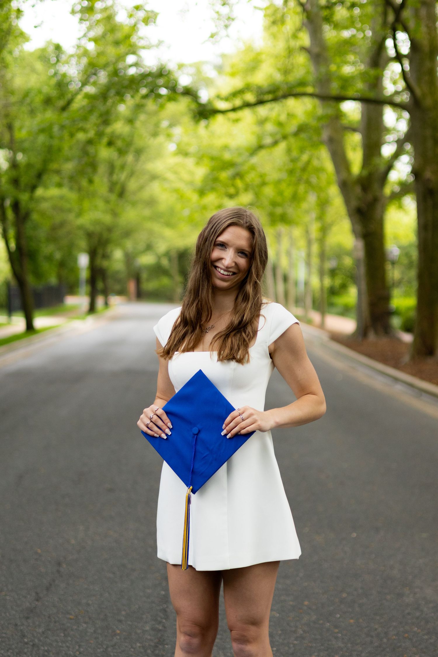 Person in white dress holds blue graduation cap on tree-lined road at the University of Delaware.