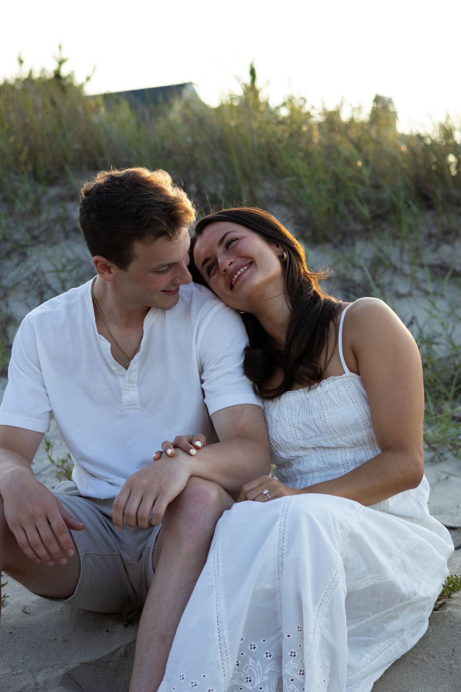 Couple in white outfits share intimate moments on a sandy beach at sunset during engagement photos in Lewes Beach, Delaware.