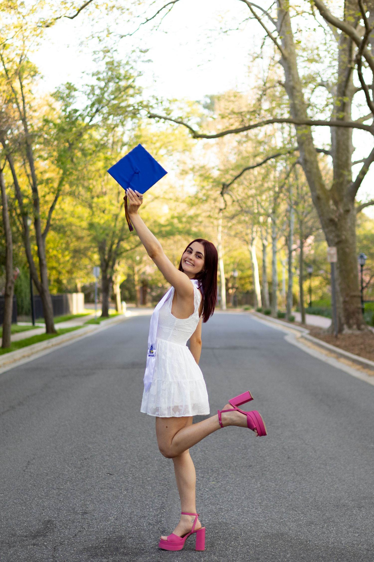 Celebratory graduation photos with blue graduation cap and pink heels on a tree-lined street at the University of Delaware.