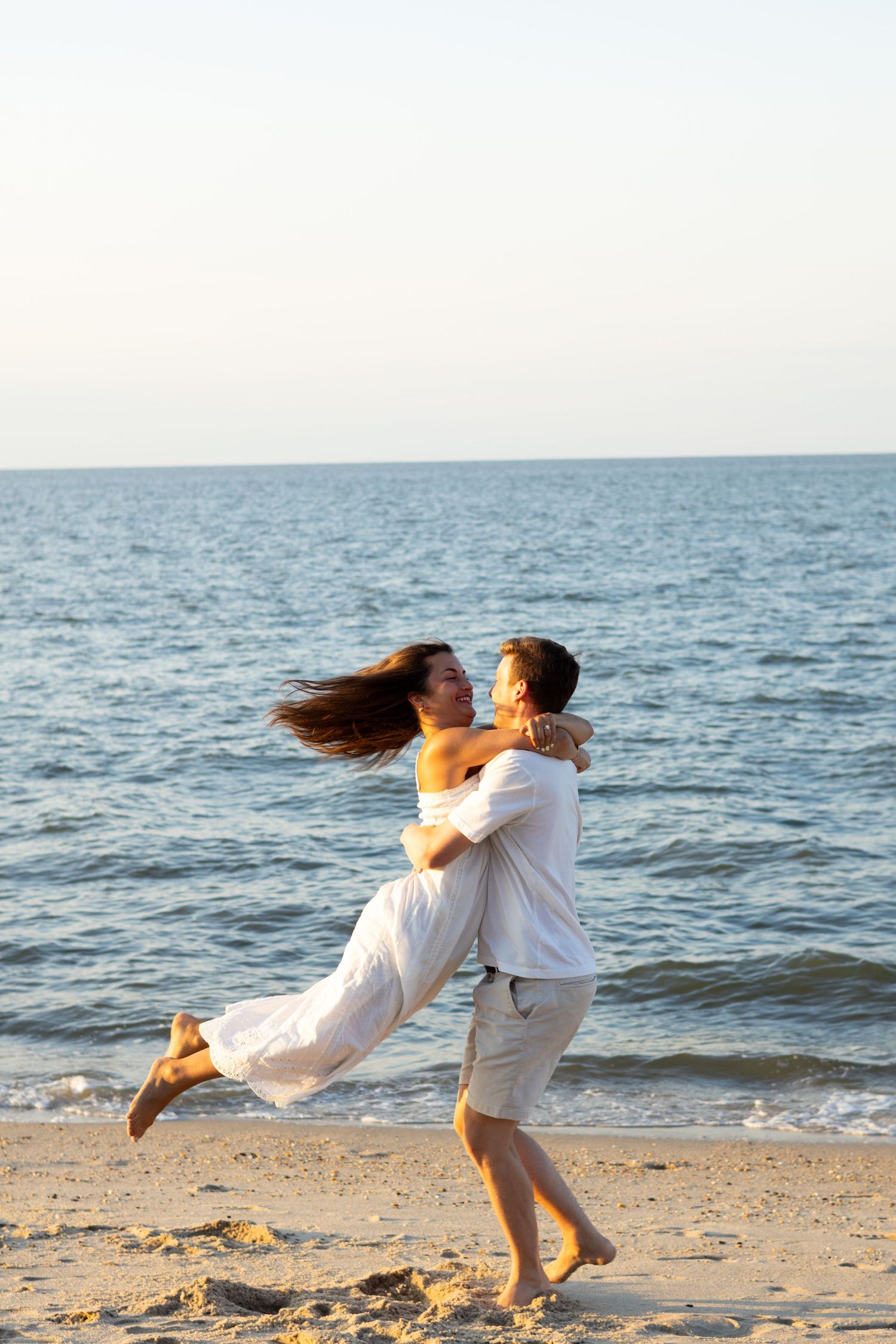 Playful couple in white clothing spinning and dancing on beach at sunset during engagement photos in Lewes, Delaware.