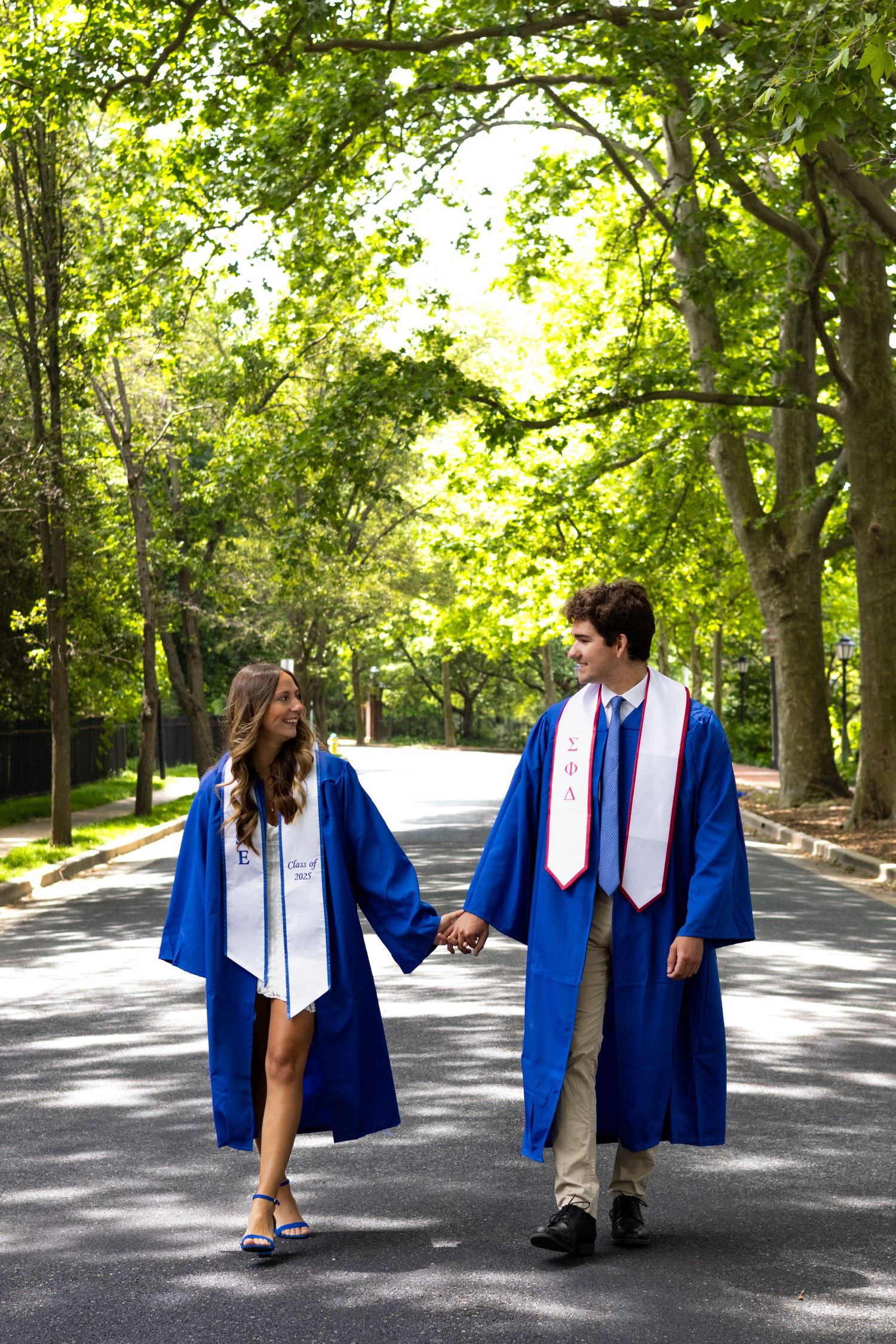 Graduates walking together on tree-lined campus path in blue academic regalia at the Univeristy of Delaware.