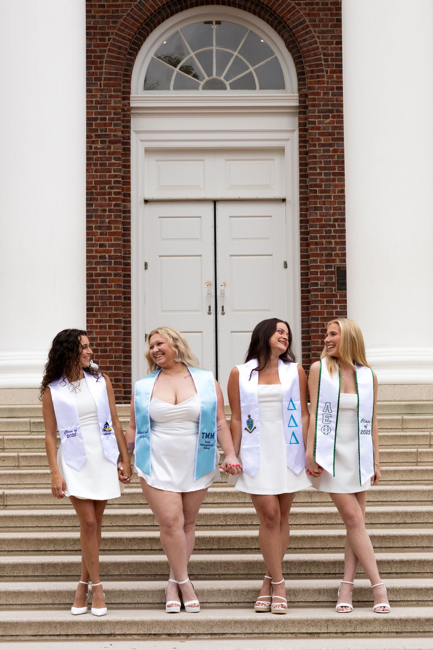 Group wearing white dresses and graduation sashes pose on university steps at the University of Delaware.