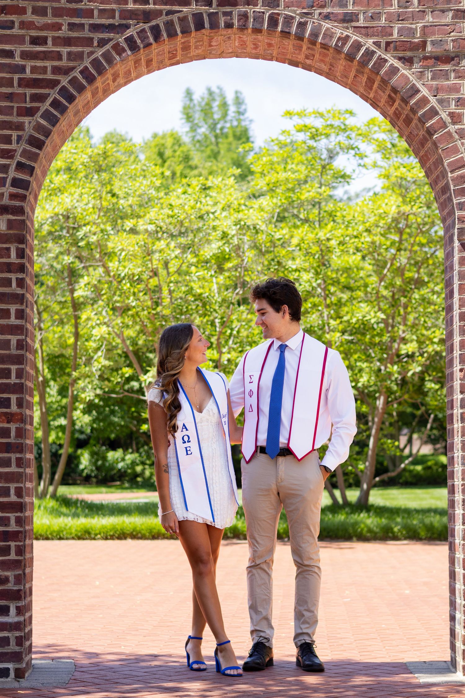 Couple in graduation attire walking under brick archway on sunny campus day at the University of Delaware.