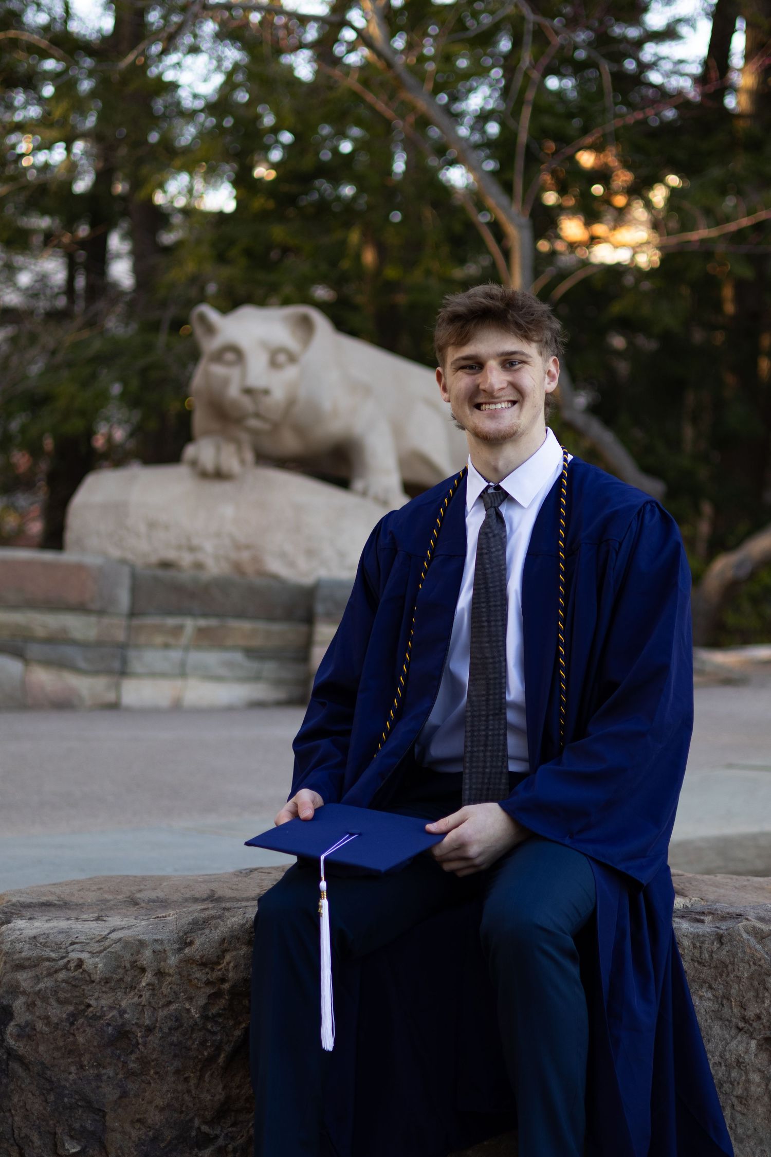 Graduate in blue cap and gown sits on stone wall at sunset at Penn State University.