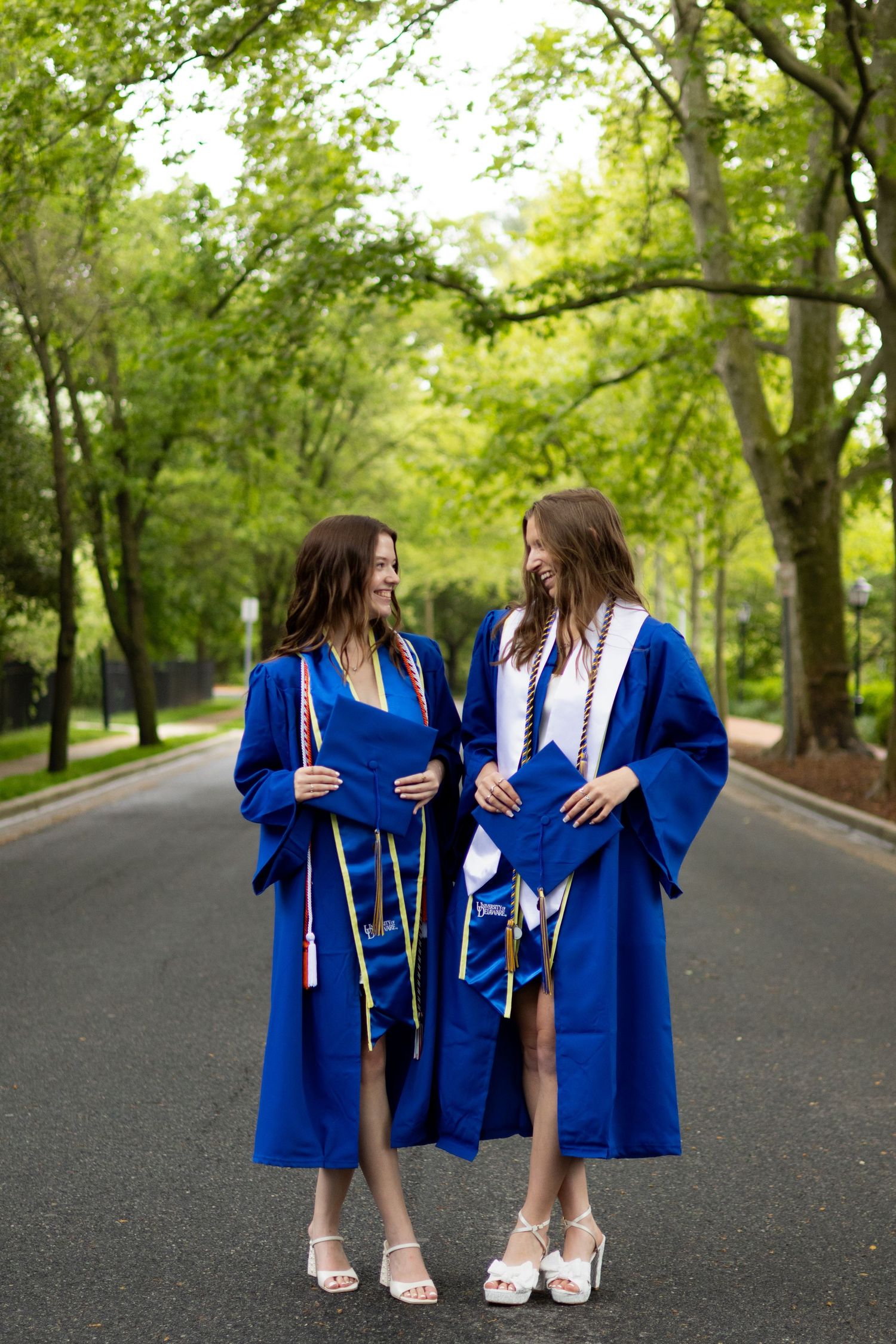 Two graduates in blue robes share a joyful moment on a tree-lined path at the University of Delaware.