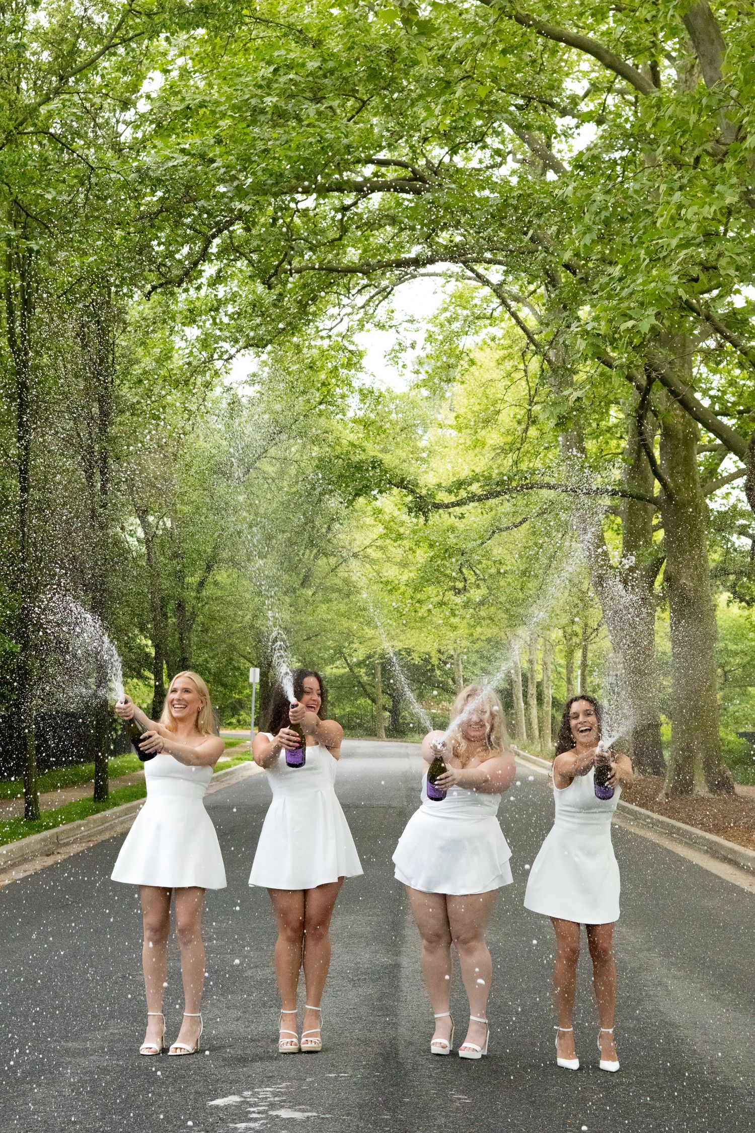 Graduates in white dresses celebrate with champagne spray on tree-lined path at the University of Delaware.