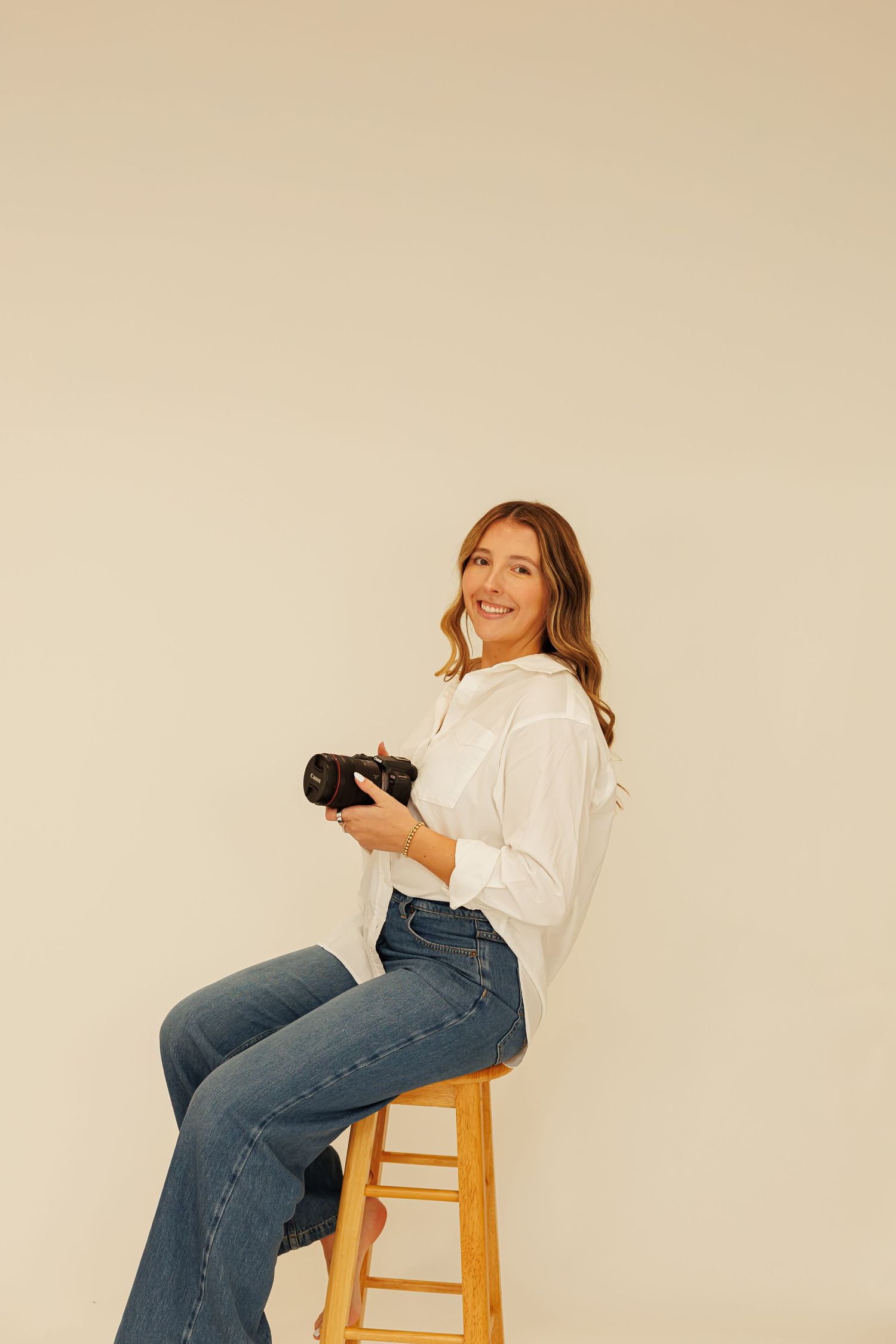 Lifestyle portrait of photographer with camera sitting on wooden stool against beige background for Madison Adele Photography.